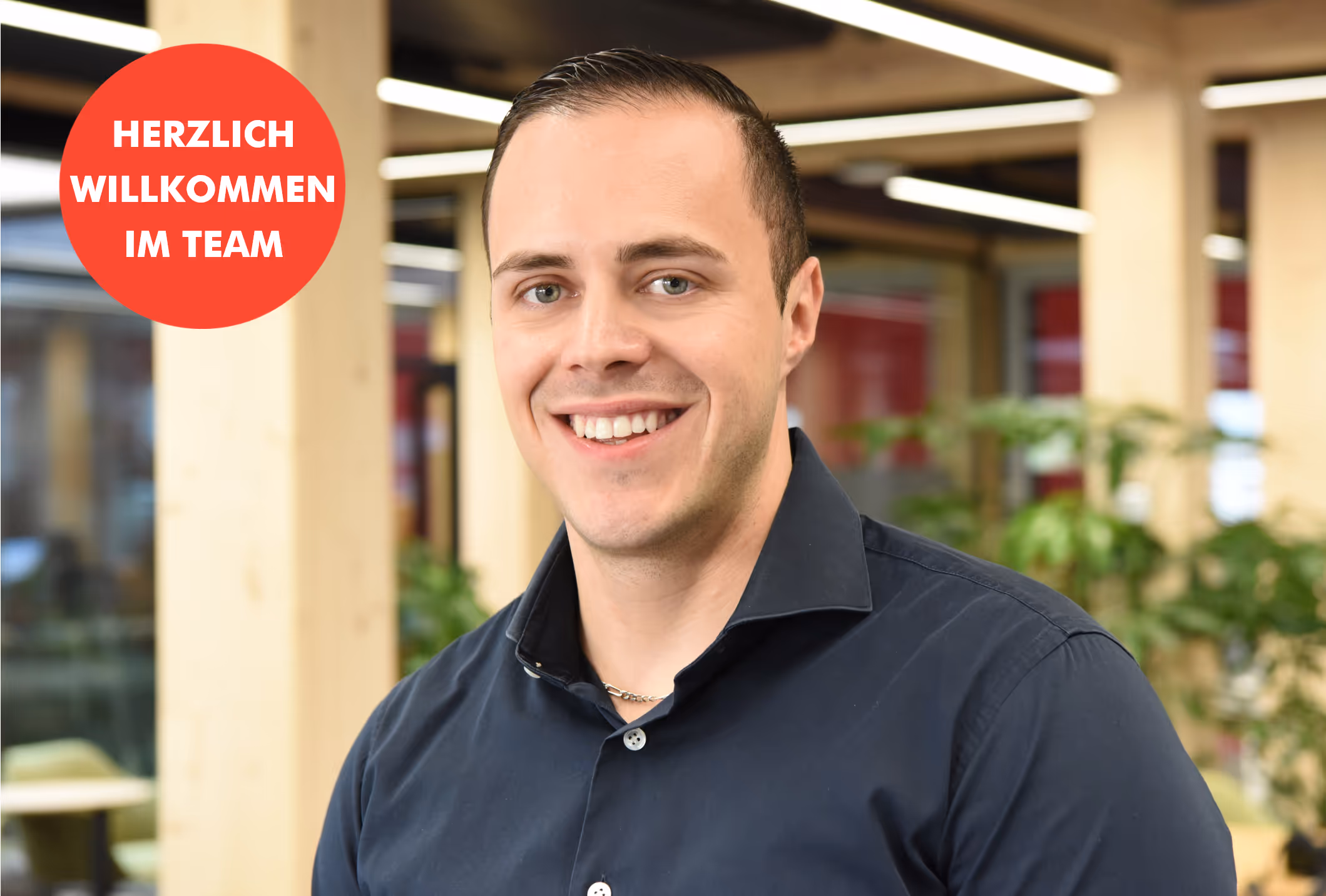 Lächelnder Mann im dunklen Hemd im Büro mit Begrüssungsbutton im Team.