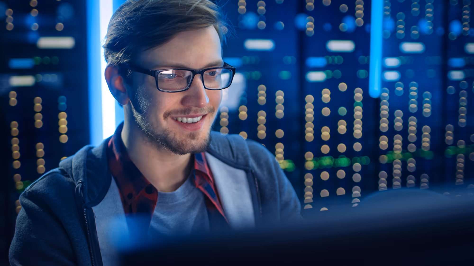 Smiling man with glasses works at a screen in front of lit server racks in a data centre.
