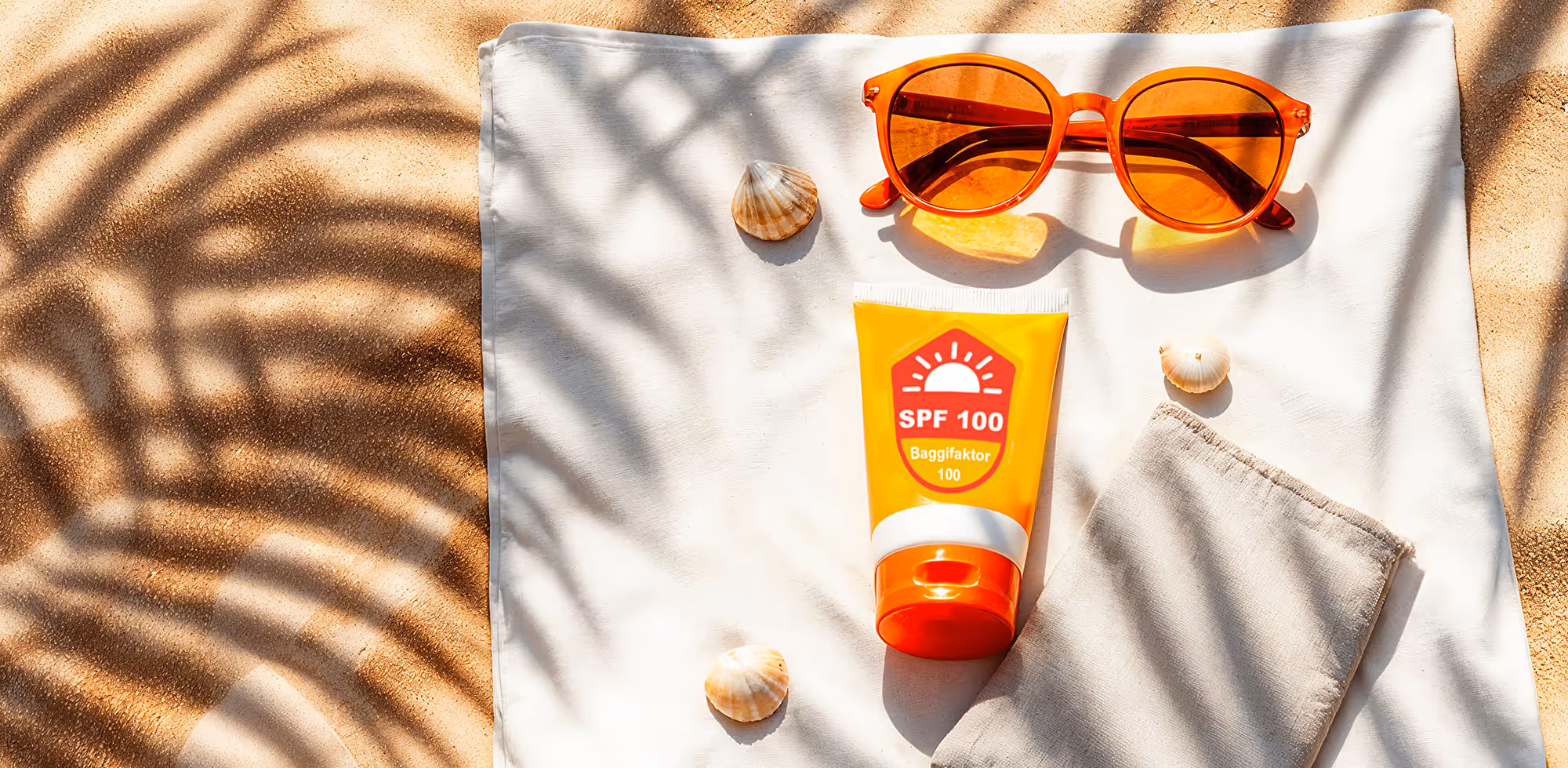 Sunscreen with SPF 100 and orange sunglasses on a cloth in the sand with palm-leaf shadows.