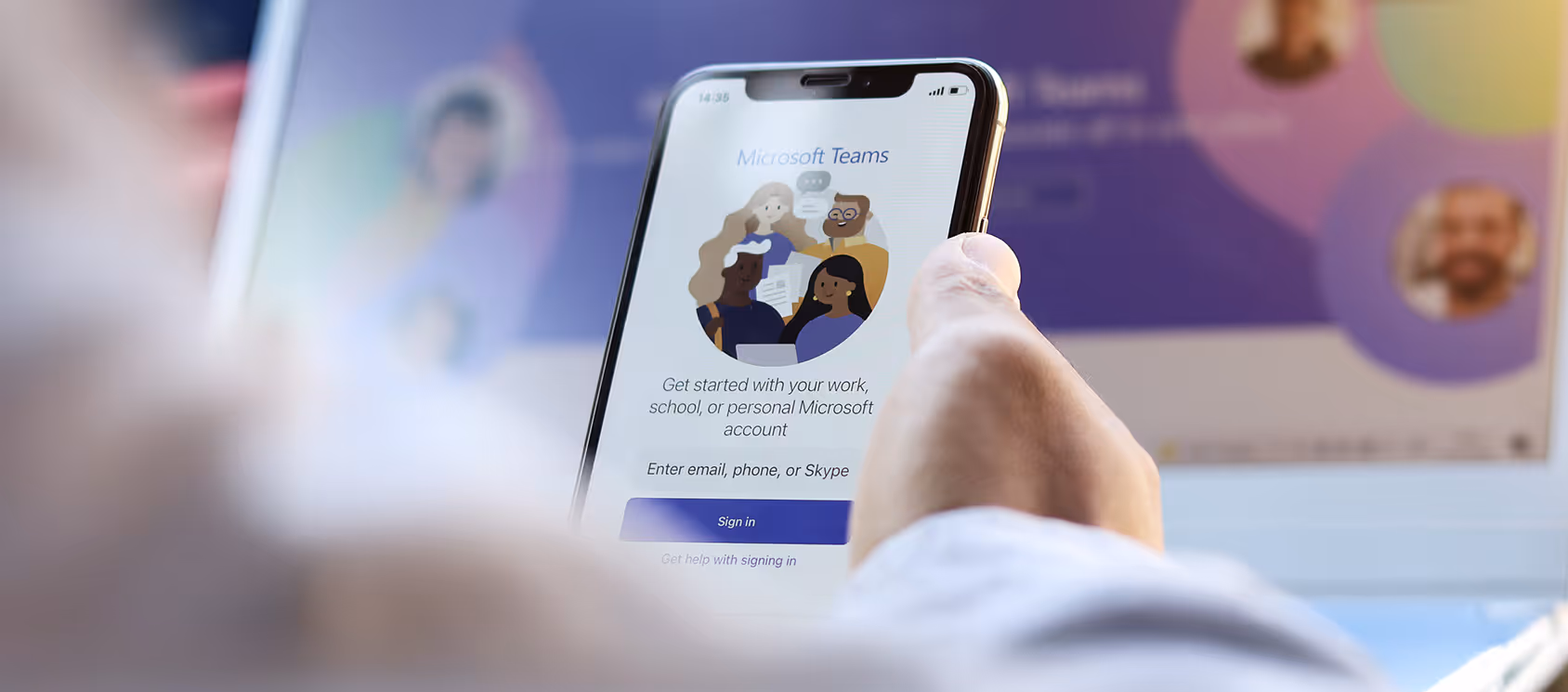 Hand holding a smartphone showing the Microsoft Teams sign-in screen, a laptop in the background.