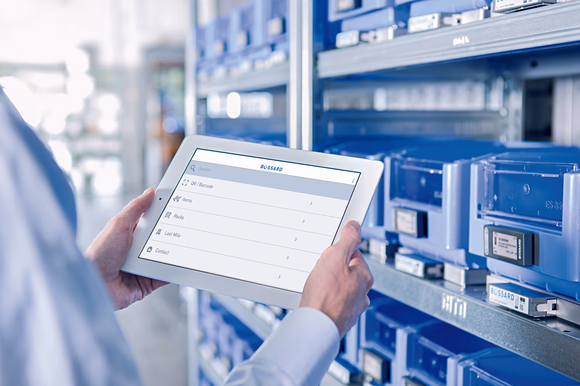 Hand hält ein Tablet mit Bossard-App vor Lagerregalen voller blauer Kleinteilebehälter