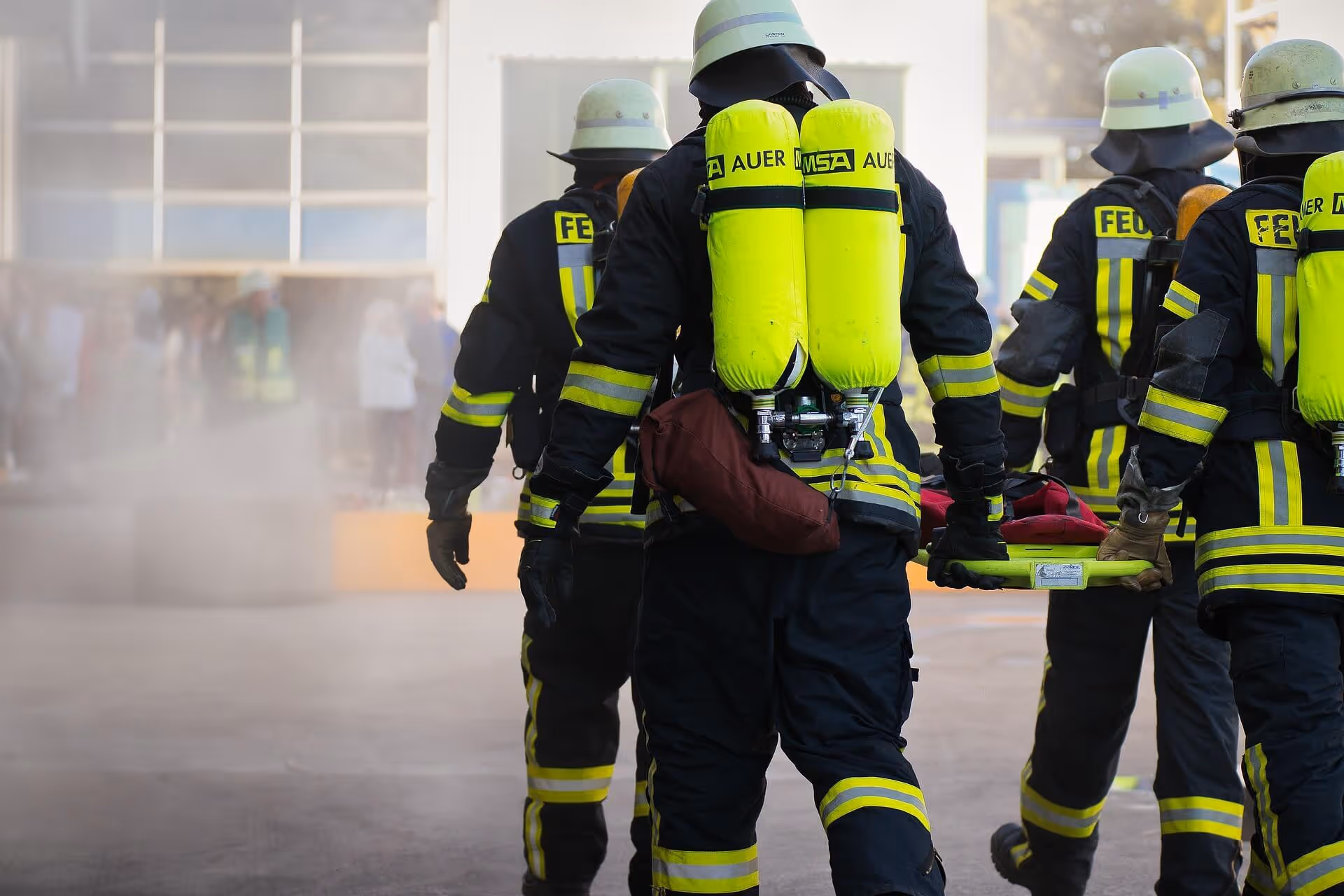 Feuerwehrleute mit Atemschutzgeräten tragen bei einer Übung eine Trage durch Rauch vor einem Gebäude.