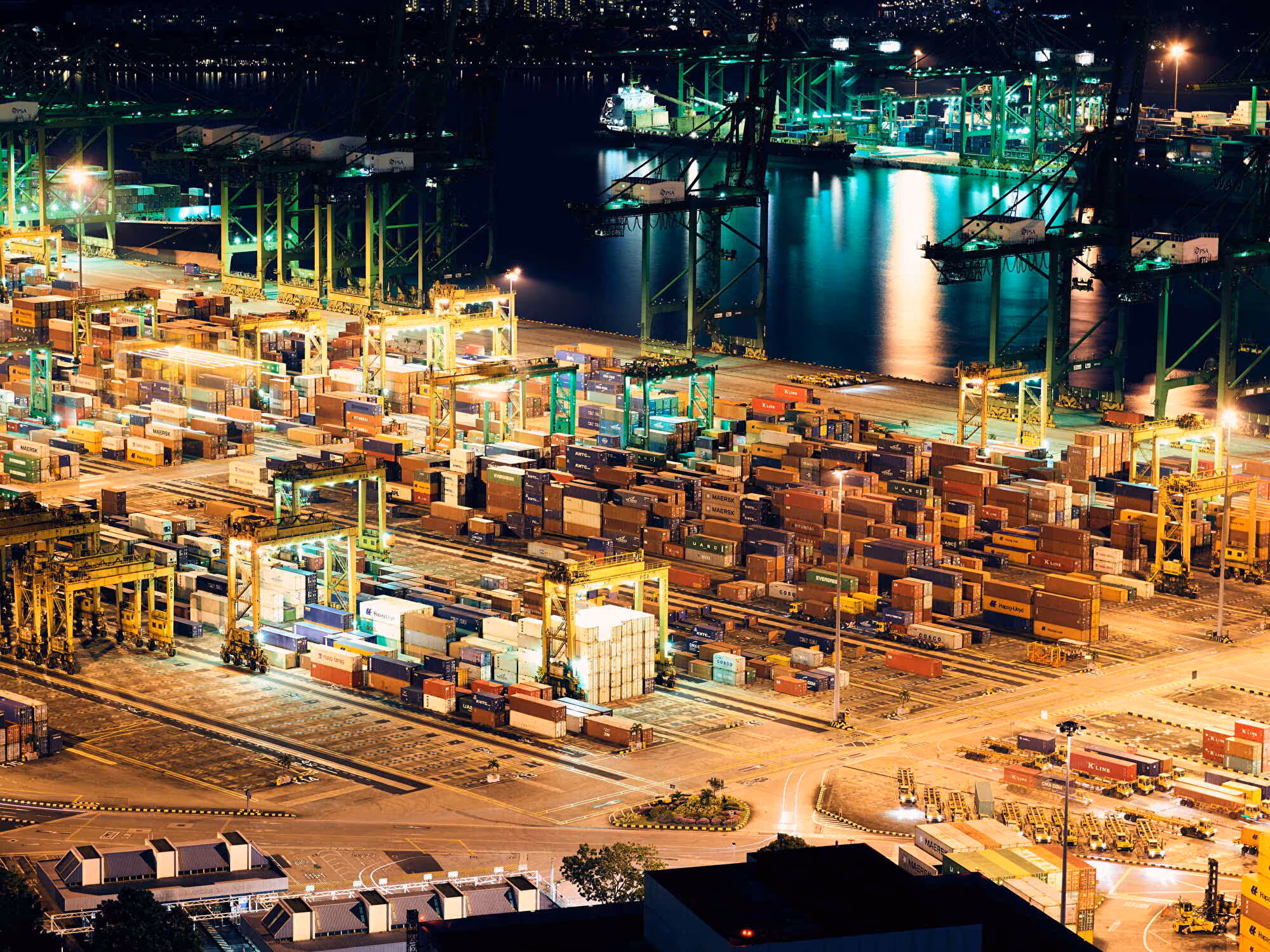 Beleuchteter Containerhafen bei Nacht mit Kränen, gestapelten Frachtcontainern und Schiffen am Wasser.