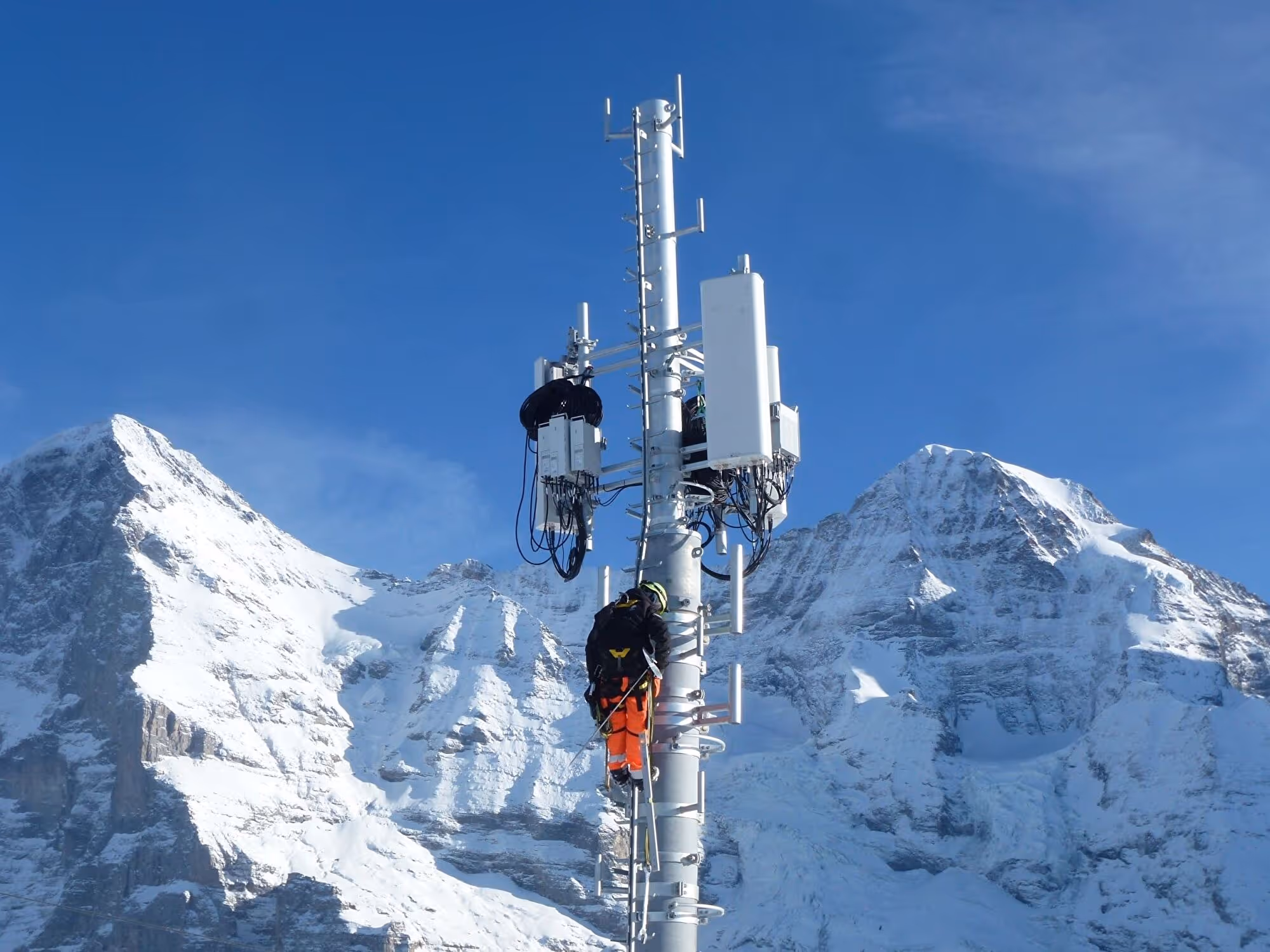 Zwei Techniker montieren Antennen an einem Funkmast vor verschneiten Berggipfeln unter blauem Himmel.