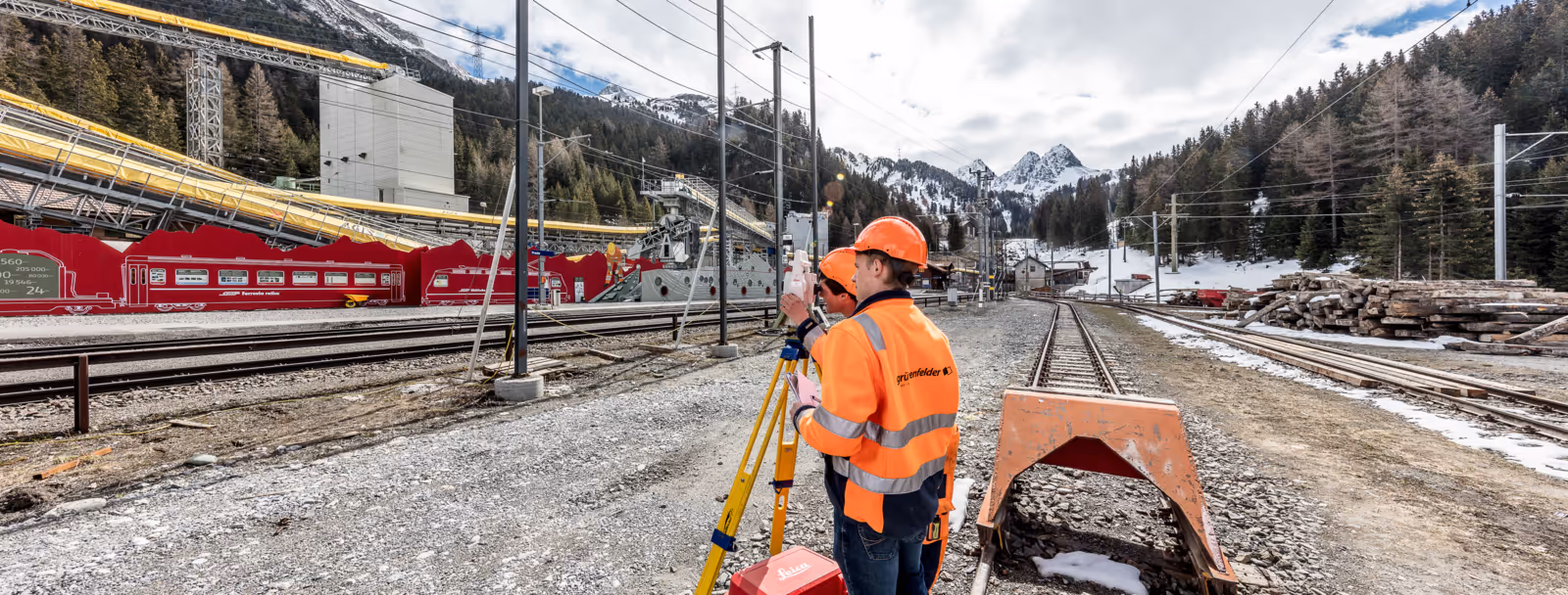 Vermesser in oranger Warnkleidung an einem Bahngleis vor verschneiter Bergkulisse