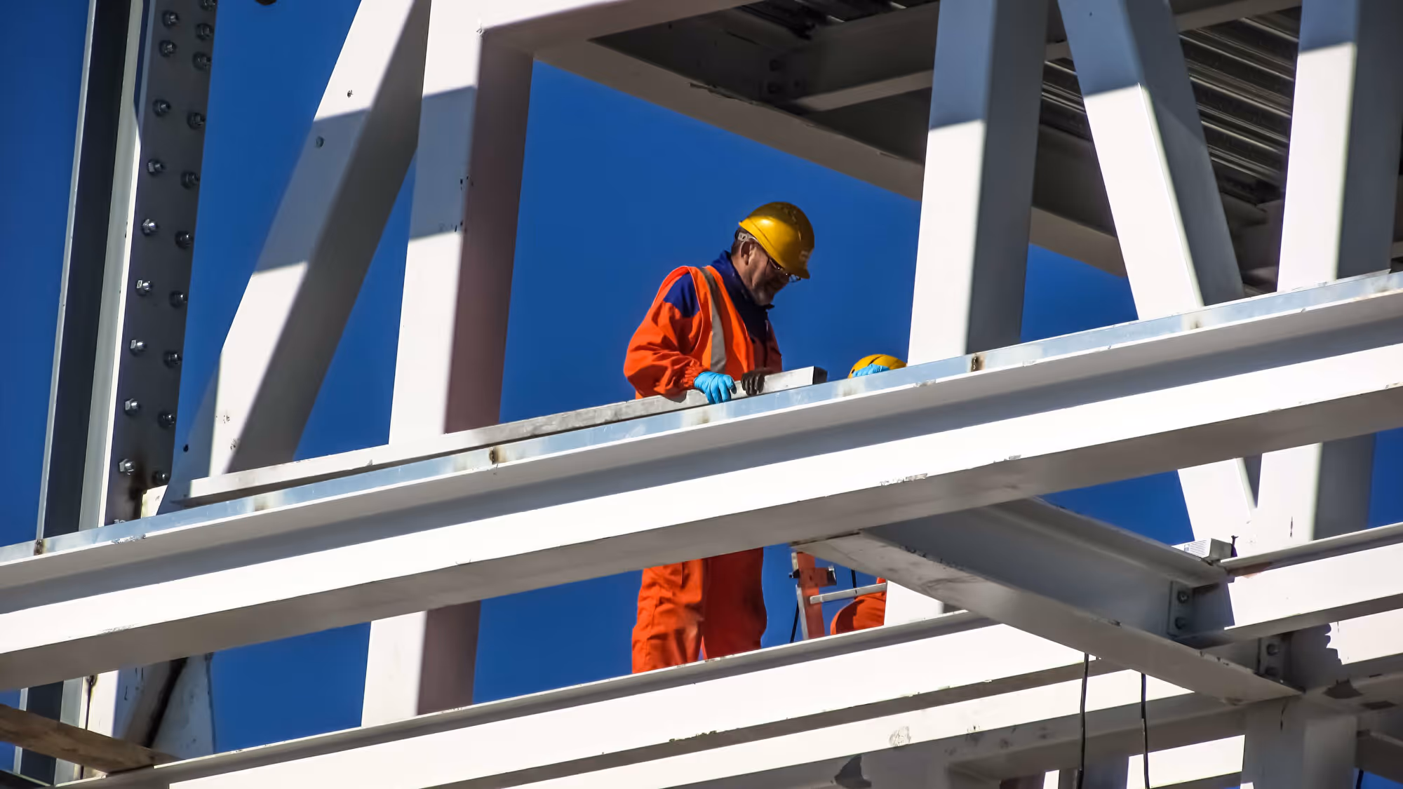 Bauarbeiter in oranger Schutzkleidung mit Helm arbeitet auf einer Stahlkonstruktion vor blauem Himmel.