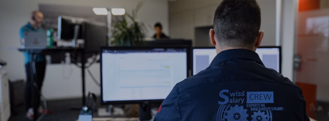 Mitarbeiter von hinten am Bildschirm, Jacke mit Aufdruck SwissSalary Crew im Büro.