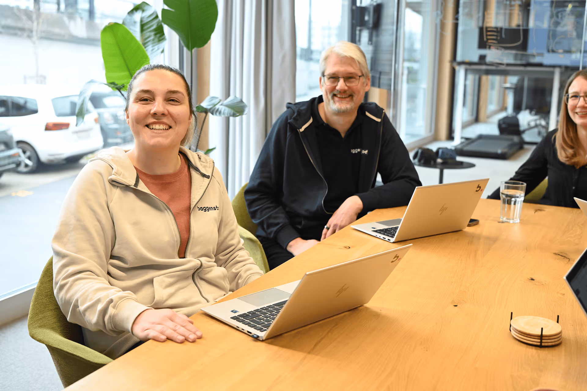 Drei lächelnde Personen sitzen mit Laptops am Holztisch im modernen Büro mit grosser Zimmerpflanze.