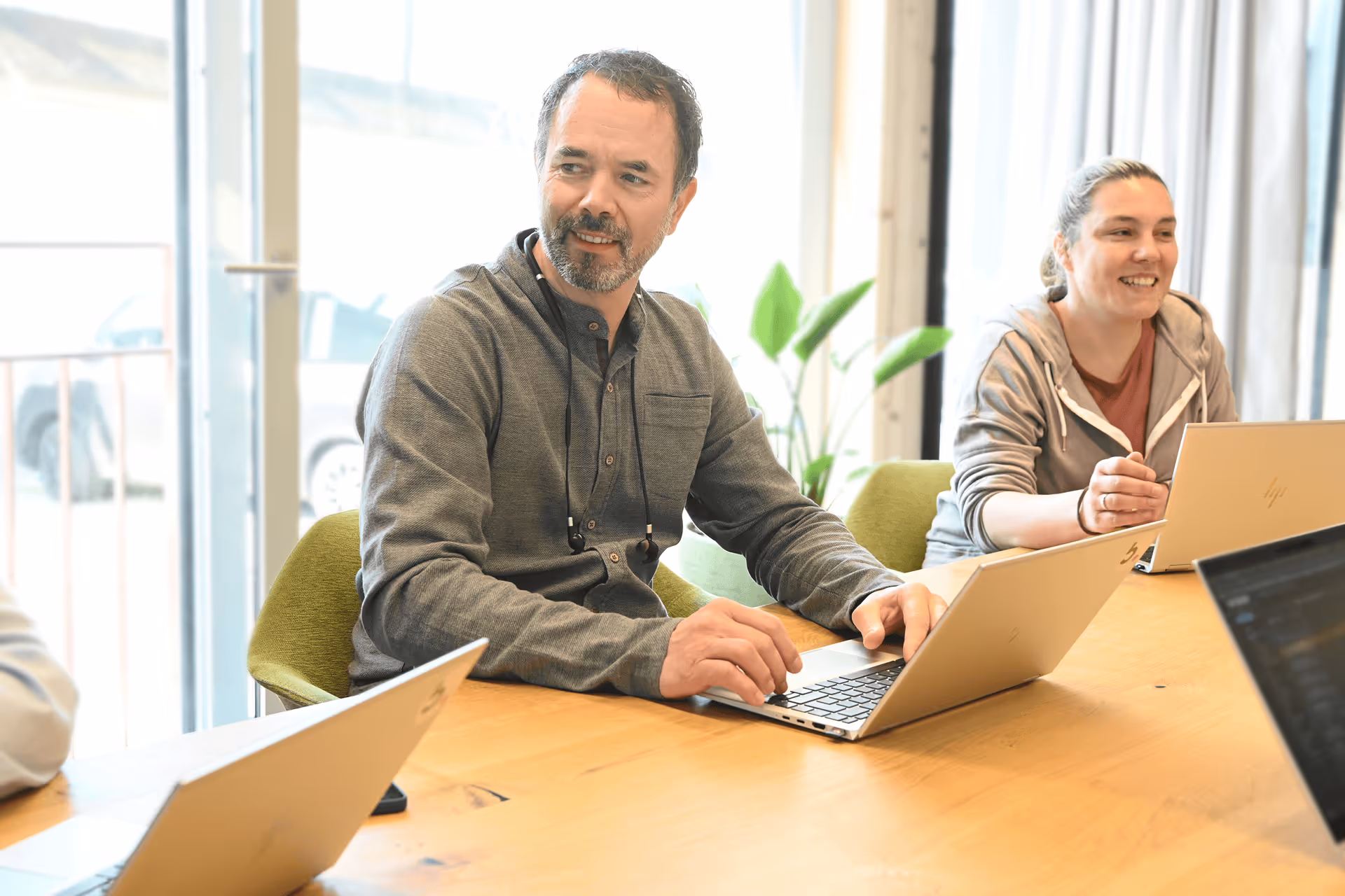 Mann mit Bart arbeitet lachend am Laptop, im Hintergrund eine Kollegin am Tisch.