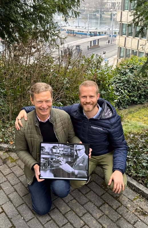 Zwei lächelnde Männer knien im Freien und halten ein gerahmtes historisches Schwarz-Weiss-Foto.