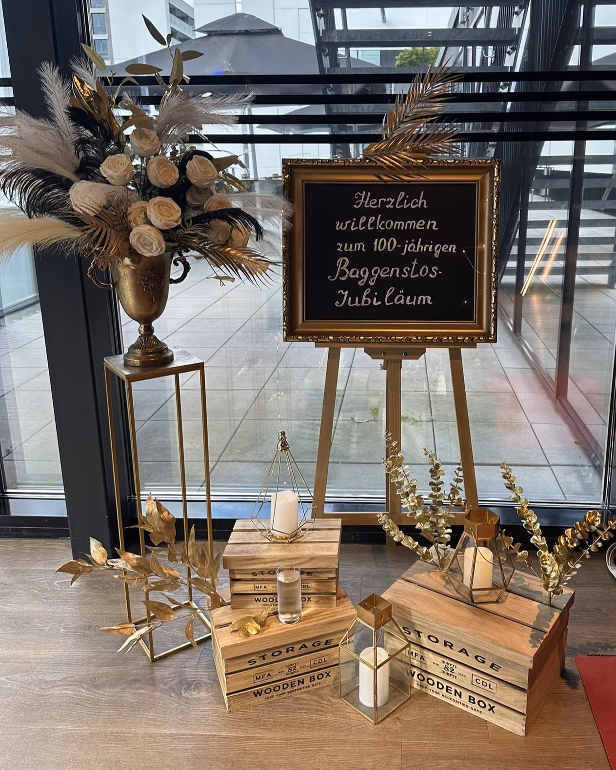 Goldene Empfangsdeko mit Tafel: Herzlich willkommen zum 100-jährigen Baggenstos-Jubiläum, neben Federstrauss.