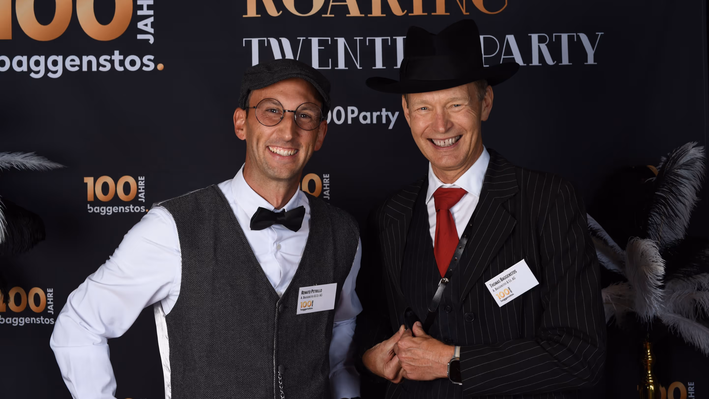 Zwei lächelnde Männer in Twenties-Kostümen vor Photowand mit 100 Jahre Baggenstos und Roaring Twenties Party.