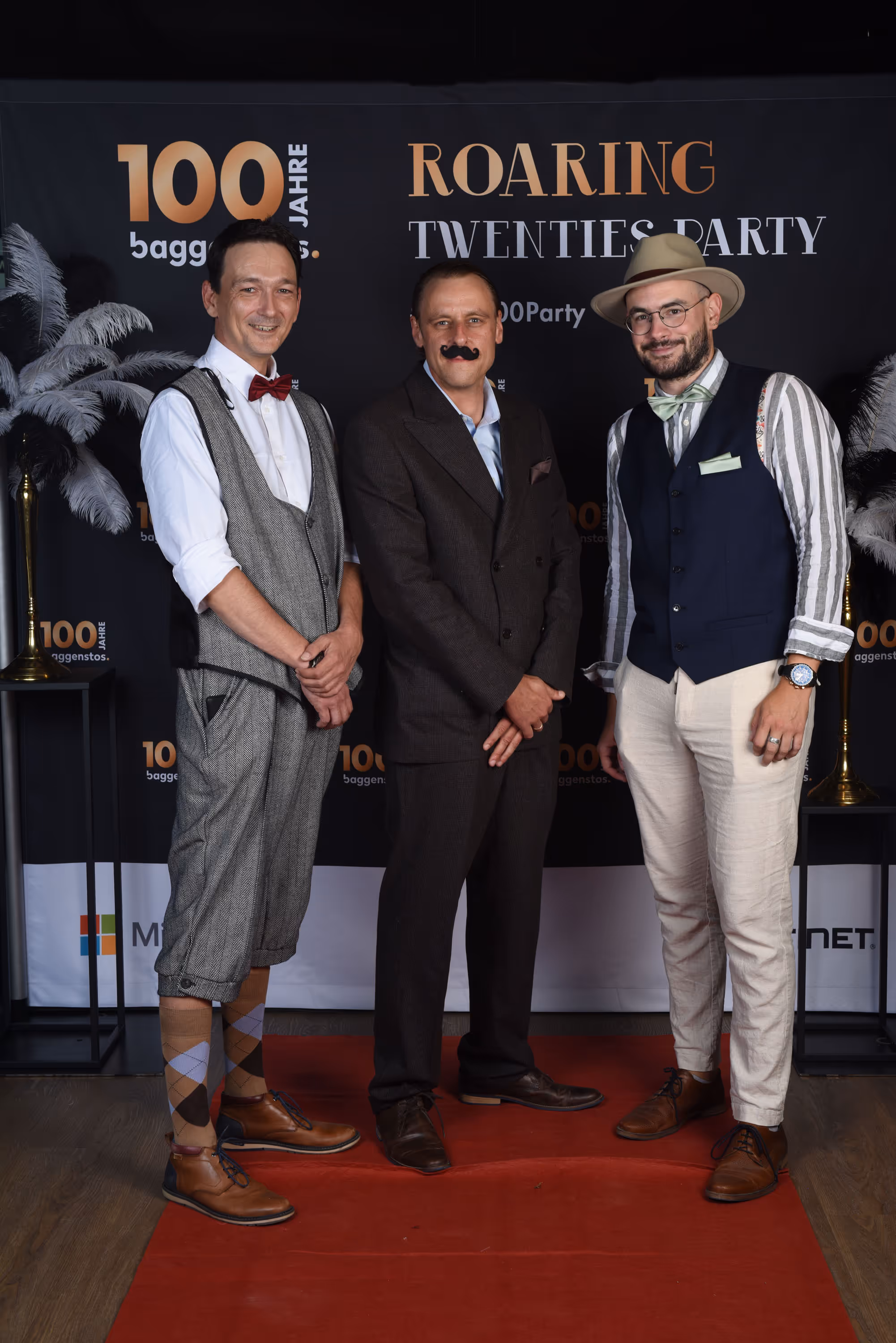 Drei Männer in eleganten 20er-Jahre-Anzügen vor Fotowand mit Aufschrift Roaring Twenties Party.