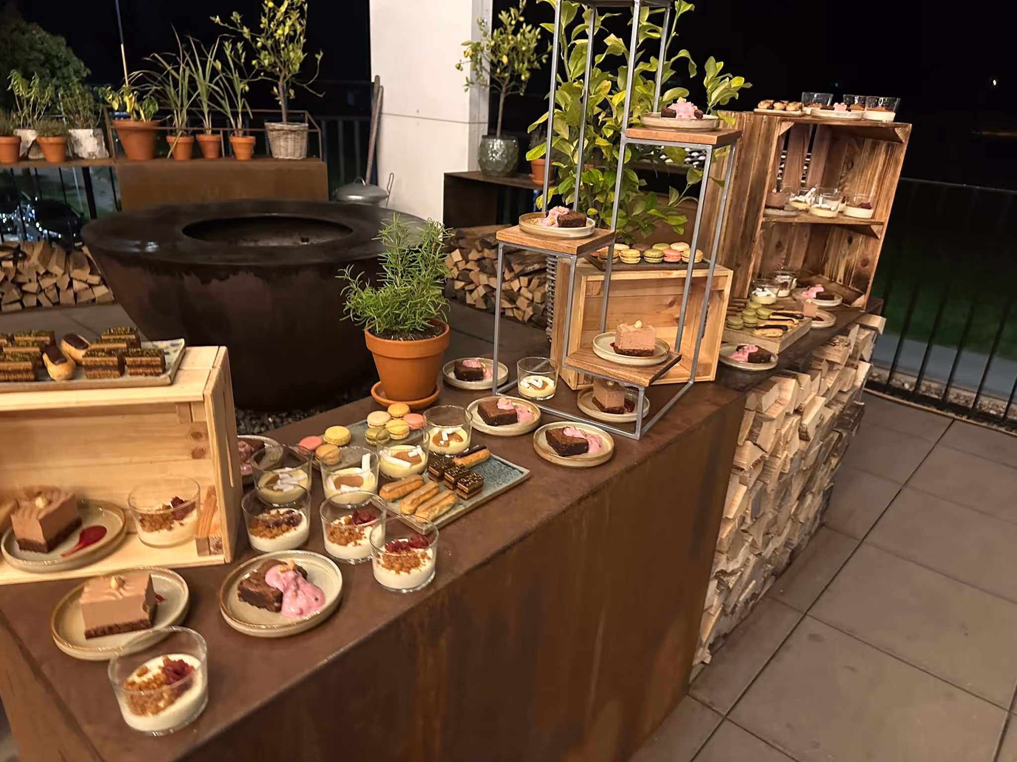 Reich gedecktes Dessertbuffet mit Kuchen und Cremes auf einer abendlichen Terrasse mit Feuerschale