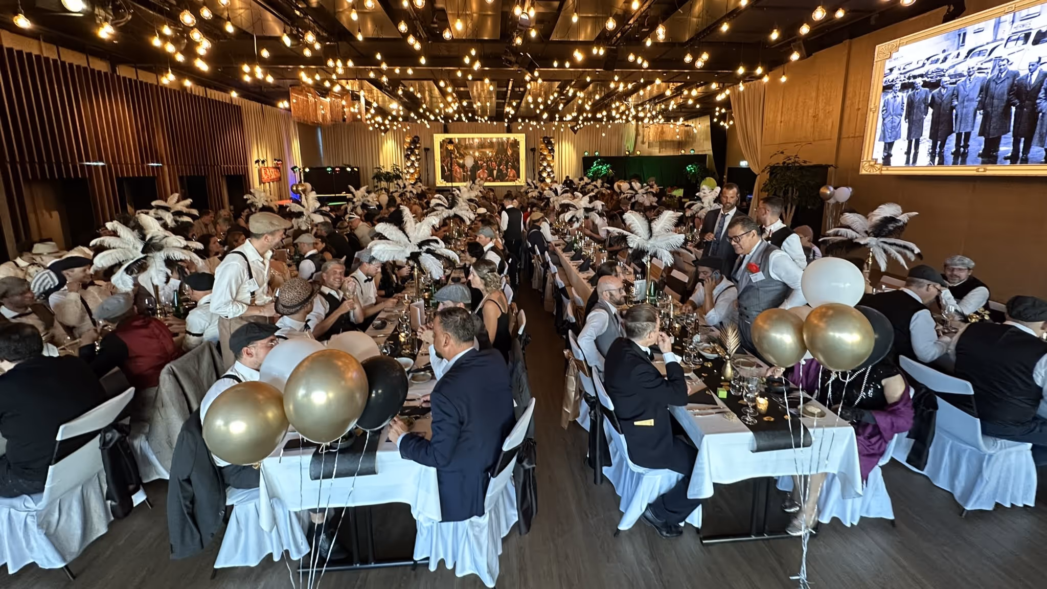 Festsaal mit vielen kostümierten Gästen an langen Tischen, Lichterketten und goldenen Ballons.
