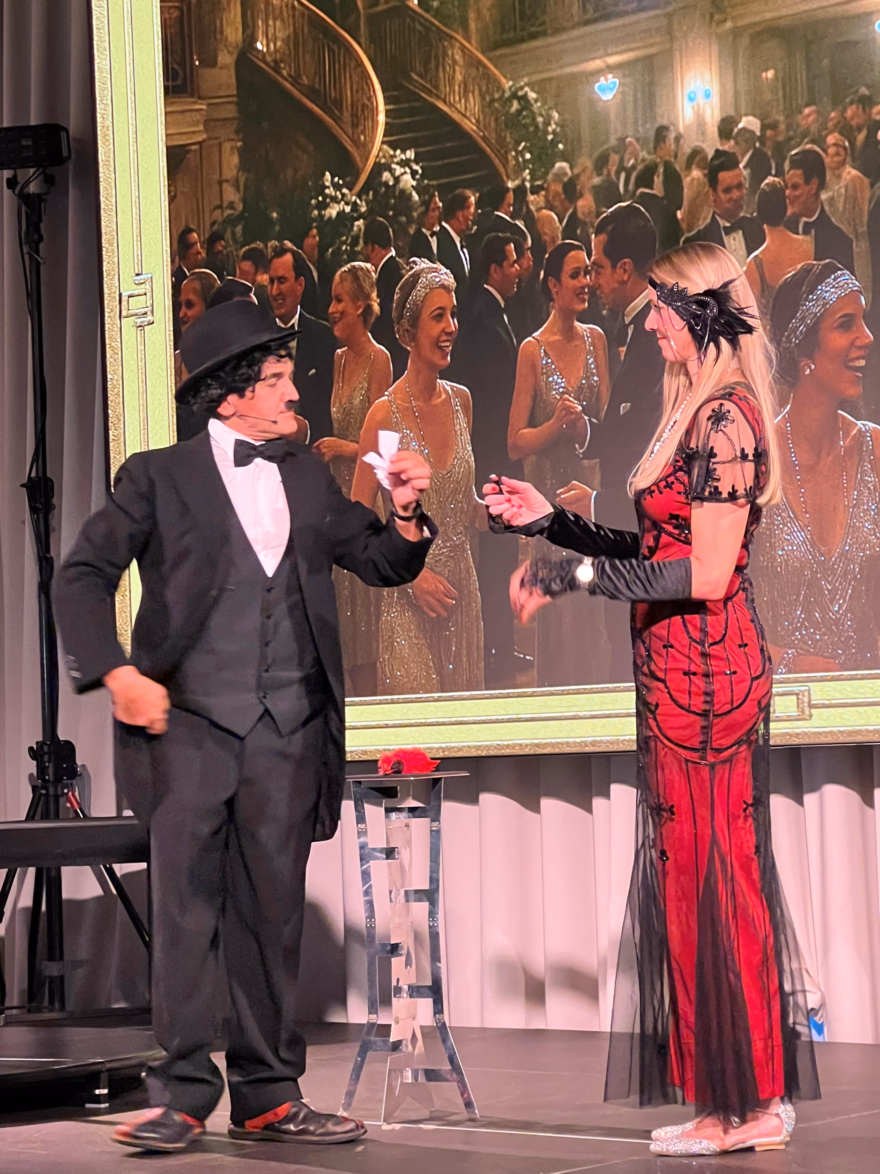 Chaplin-Darsteller und Frau im roten Kleid bei Bühnenshow vor Ballsaal-Leinwand.