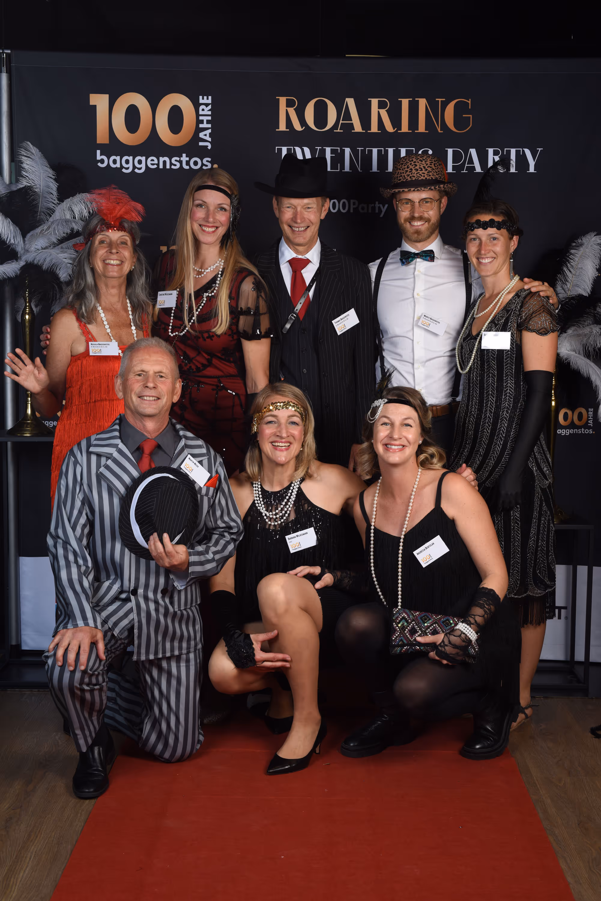 Achtköpfige Gruppe im Twenties-Look posiert vor Photowand mit 100 Jahre Baggenstos und Roaring Twenties Party.