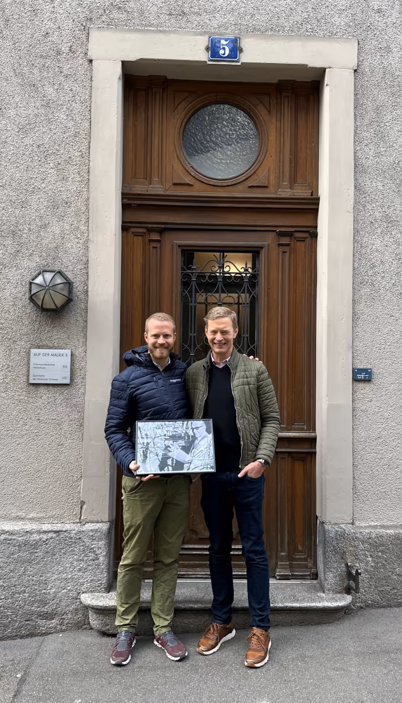 Zwei Männer stehen lächelnd mit gerahmtem Foto vor einer Holztür mit Hausnummer 5.