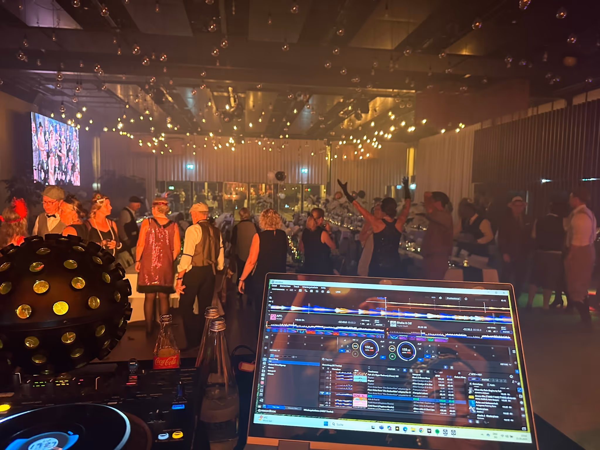 Blick vom DJ-Pult mit Laptop auf eine tanzende Menge unter funkelnden Lichtern.