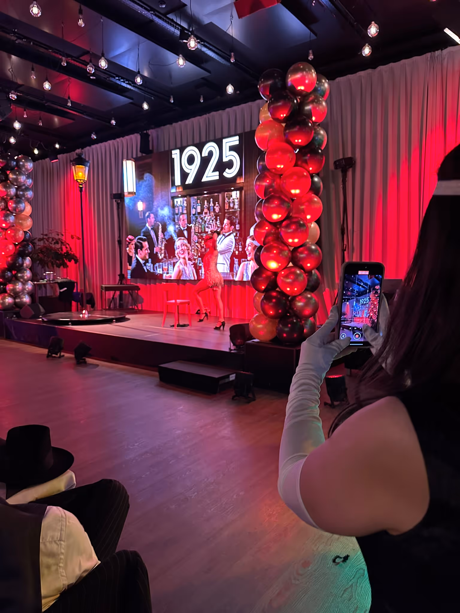 Gast filmt mit dem Smartphone eine Bühnenshow mit Leinwand, Ballonsäule und Zahl 1925.