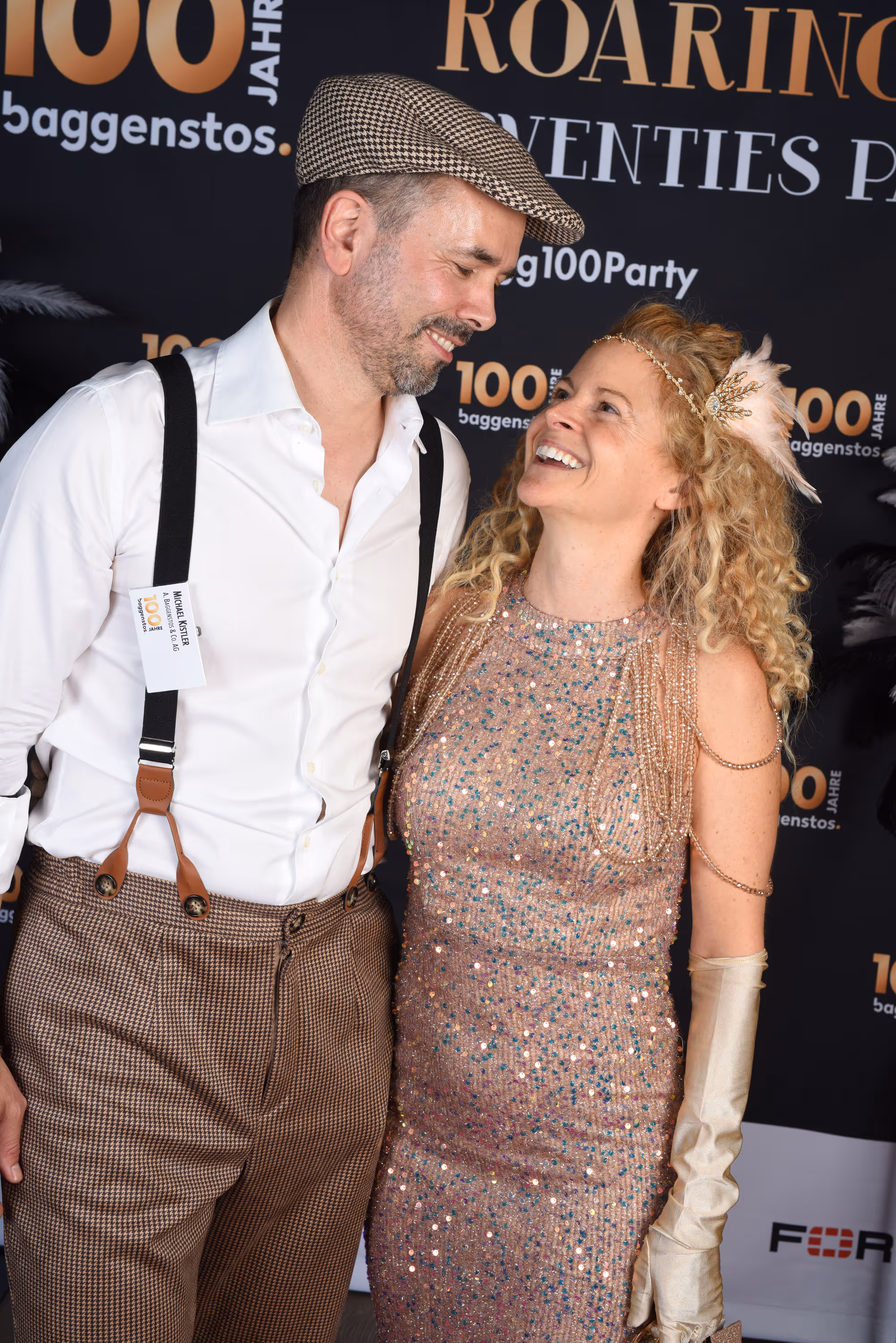 Mann mit Schiebermütze und Frau im goldenen Pailletten-Kleid lachen sich vor Fotowand an.