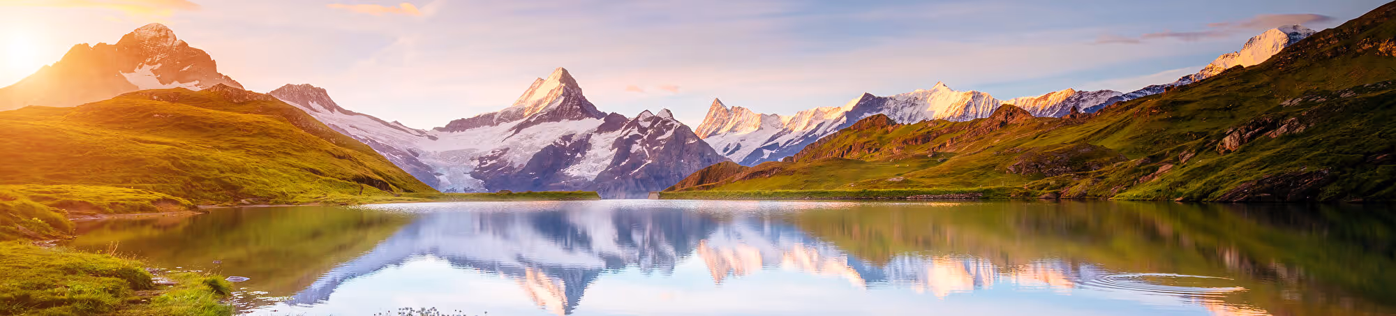 Symbolbild: schneebedeckte Schweizer Berge spiegeln sich bei Sonnenaufgang in einem ruhigen Bergsee.