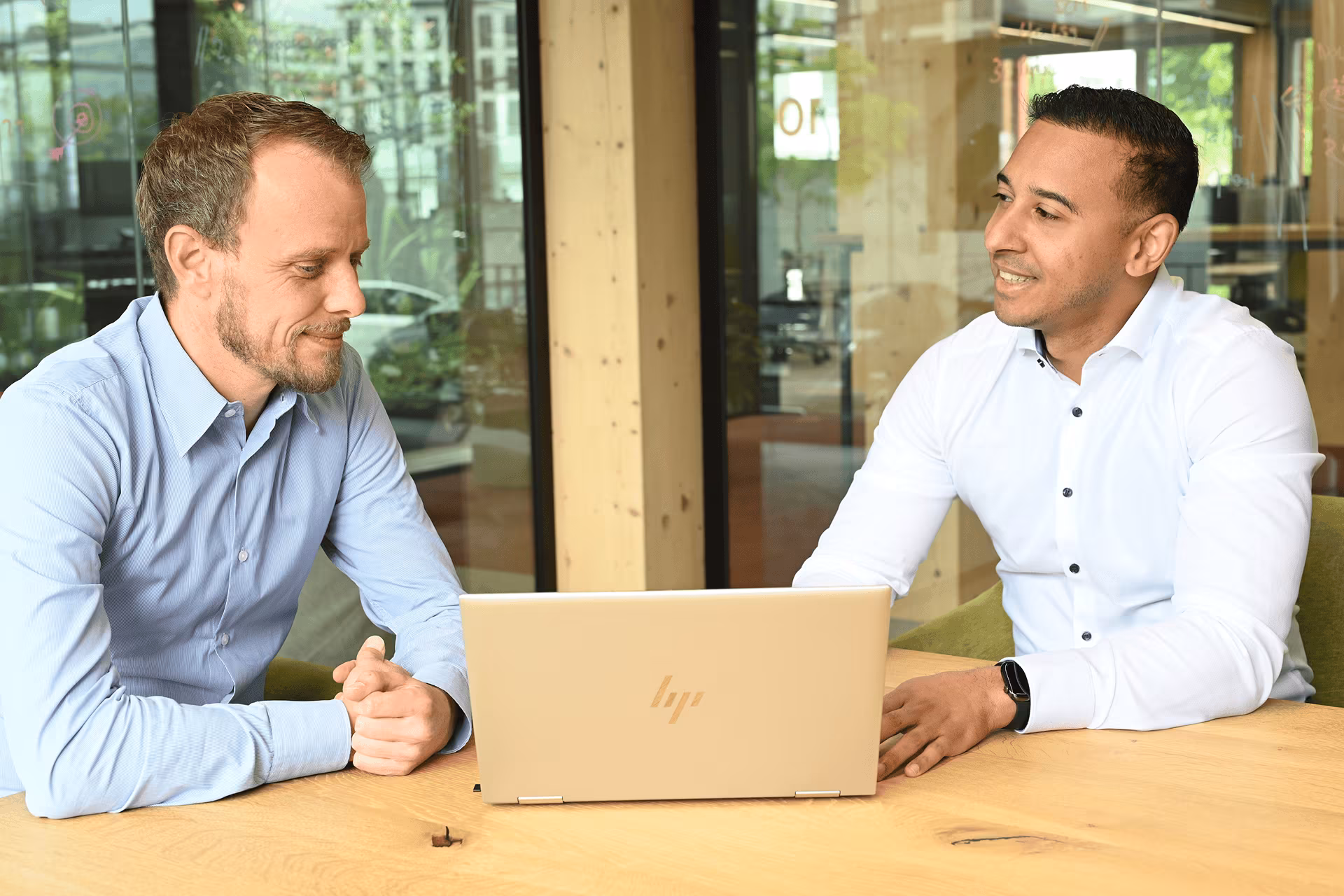 Zwei Männer sitzen an einem hellen Holztisch in einem modernen, hell beleuchteten Büro und schauen gemeinsam lächelnd auf einen geöffneten goldfarbenen HP‑Laptop zwischen ihnen; der Mann links trägt ein hellblaues Hemd, der Mann rechts ein weißes Hemd mit 