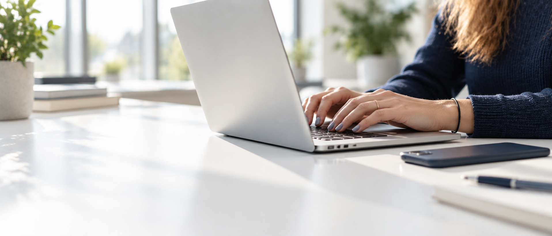 Hände einer Mitarbeiterin tippen auf einem Laptop an einem hellen Schreibtisch
