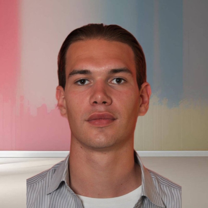 Portrait of a young man with dark hair, wearing a striped shirt over a white T-shirt, against a multicolored blurred background.