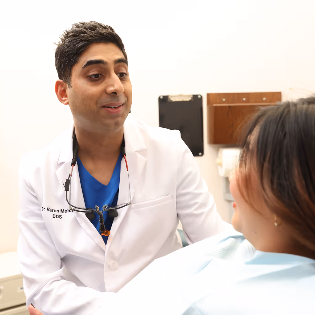 Dentist wearing a white coat and dental loupes talking to a female patient in a dental clinic.