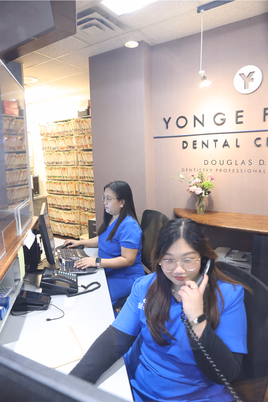 Two dental office staff members in blue scrubs working at a reception desk with computers and patient files in the background.
