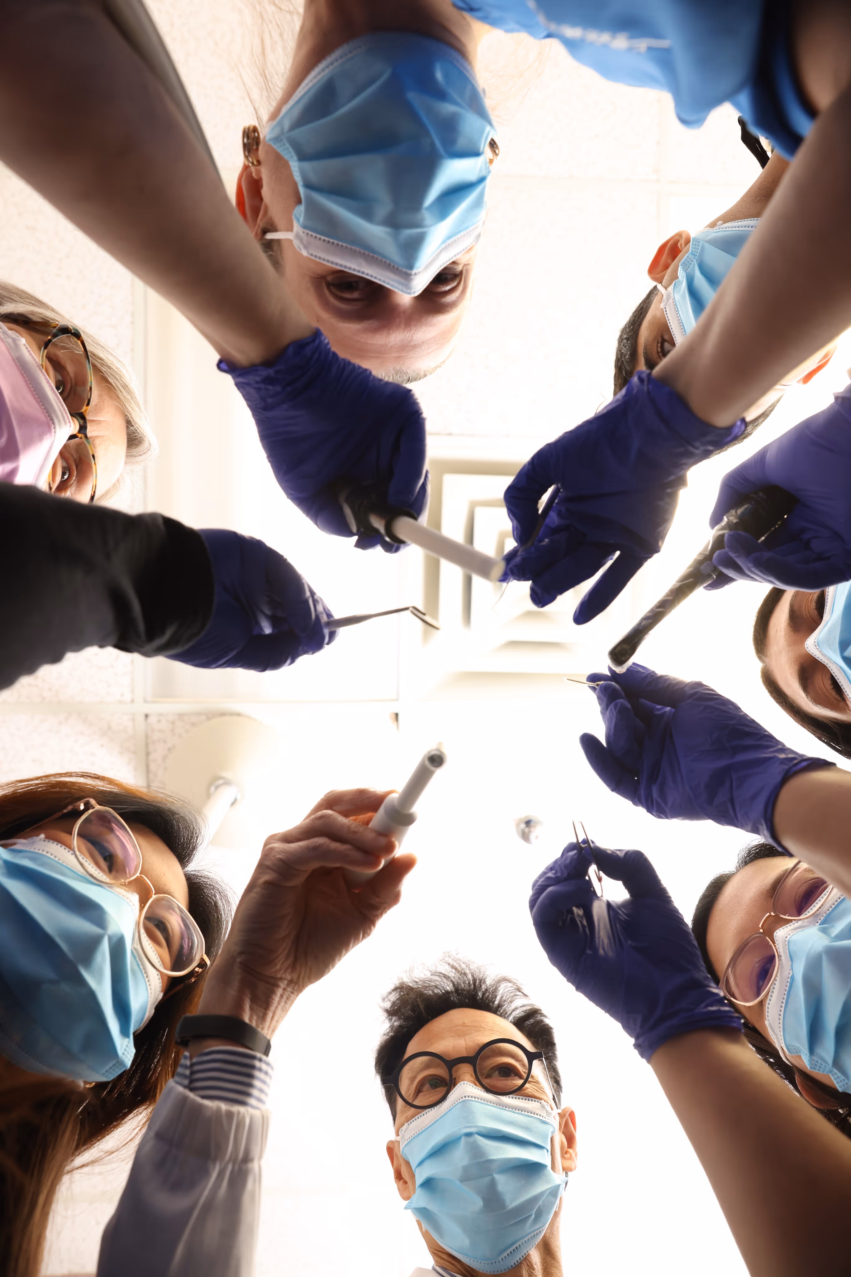 Six dental professionals wearing blue masks and gloves, holding dental tools, viewed from the patient’s perspective.