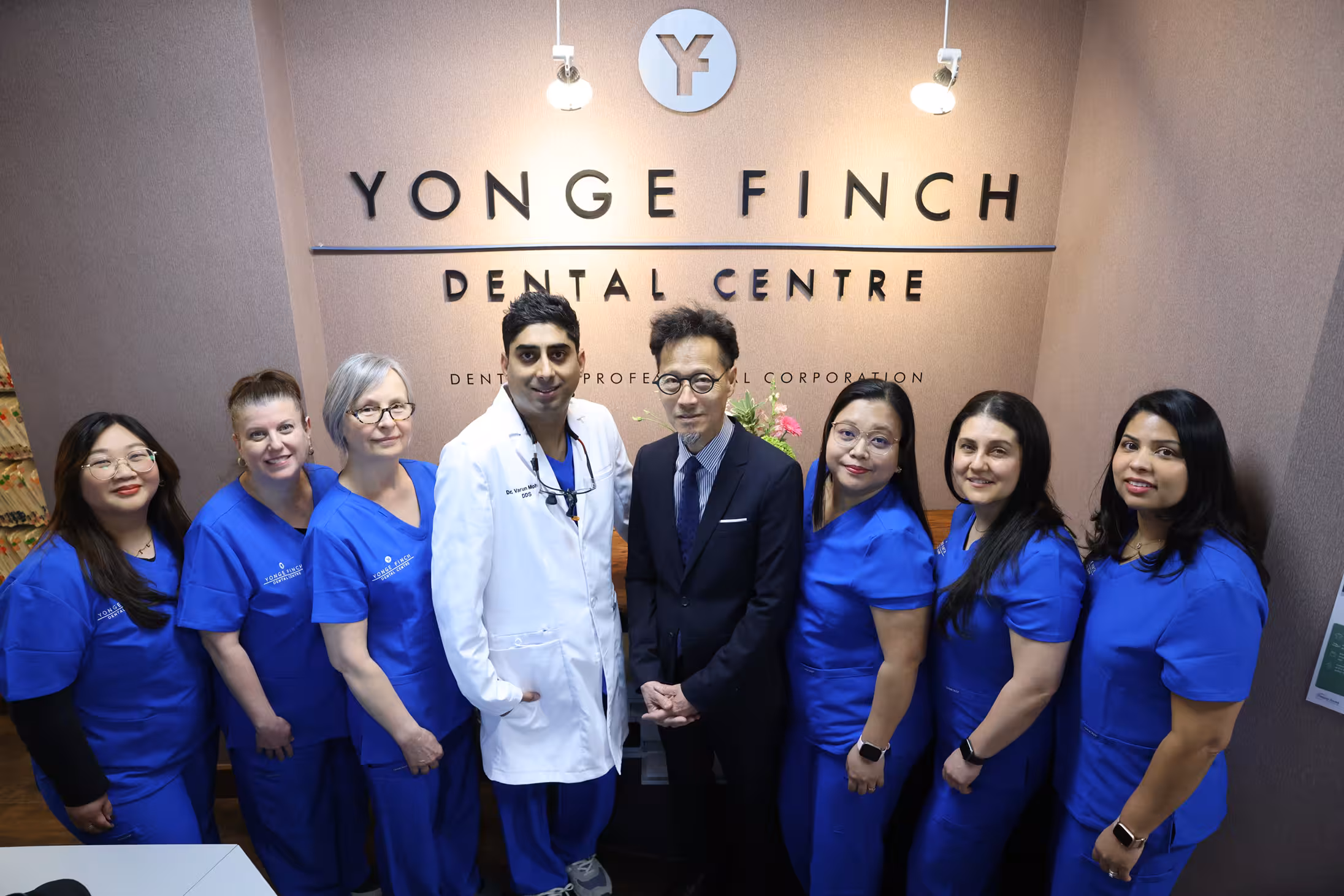 Group photo of dental professionals at Yonge Finch Dental Centre, with six women in blue scrubs, one man in a white coat, and one man in a suit standing in front of a branded wall.