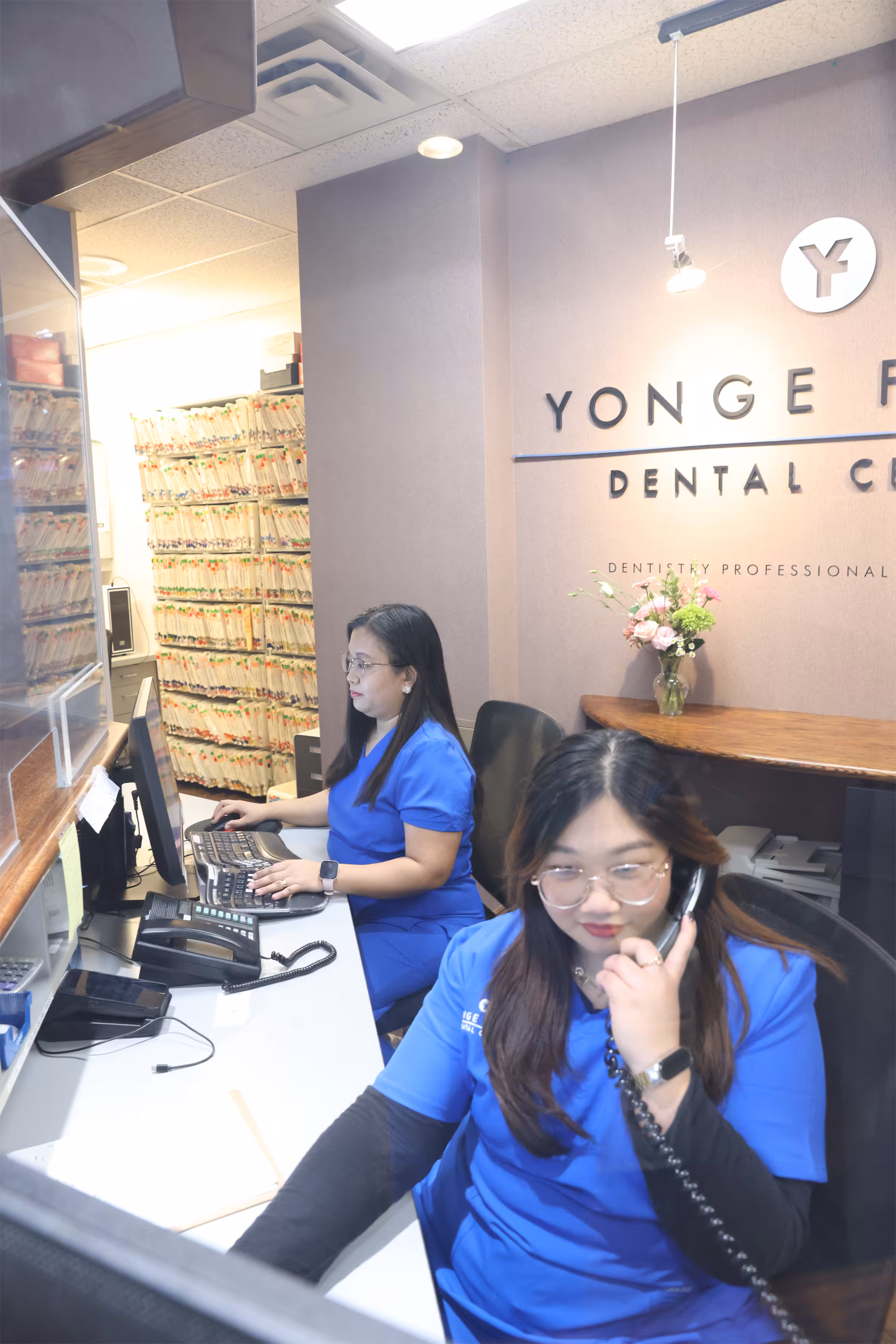 Two dental office staff in blue scrubs working at reception desks, one on the phone and the other using a computer.