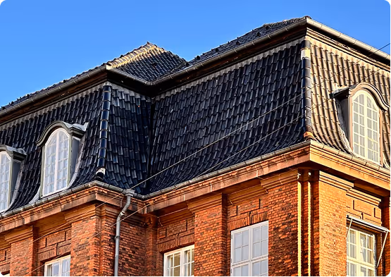 Corner view of a red brick building with a dark slate mansard roof and dormer windows under a clear blue sky.