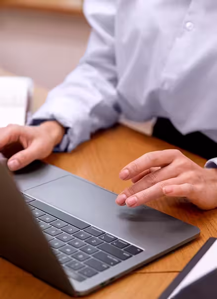 Person typing on a laptop keyboard at a wooden desk, wearing a light blue long-sleeve shirt.