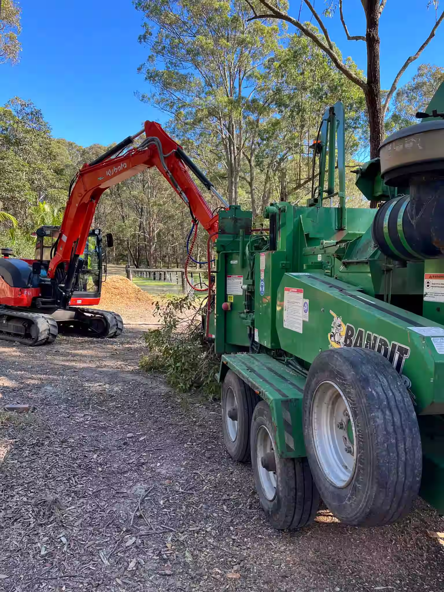 Utility worker in green helmet and orange safety gear elevated in a bucket truck working on power lines beside a rural dirt road with traffic cones and another worker on the ground.