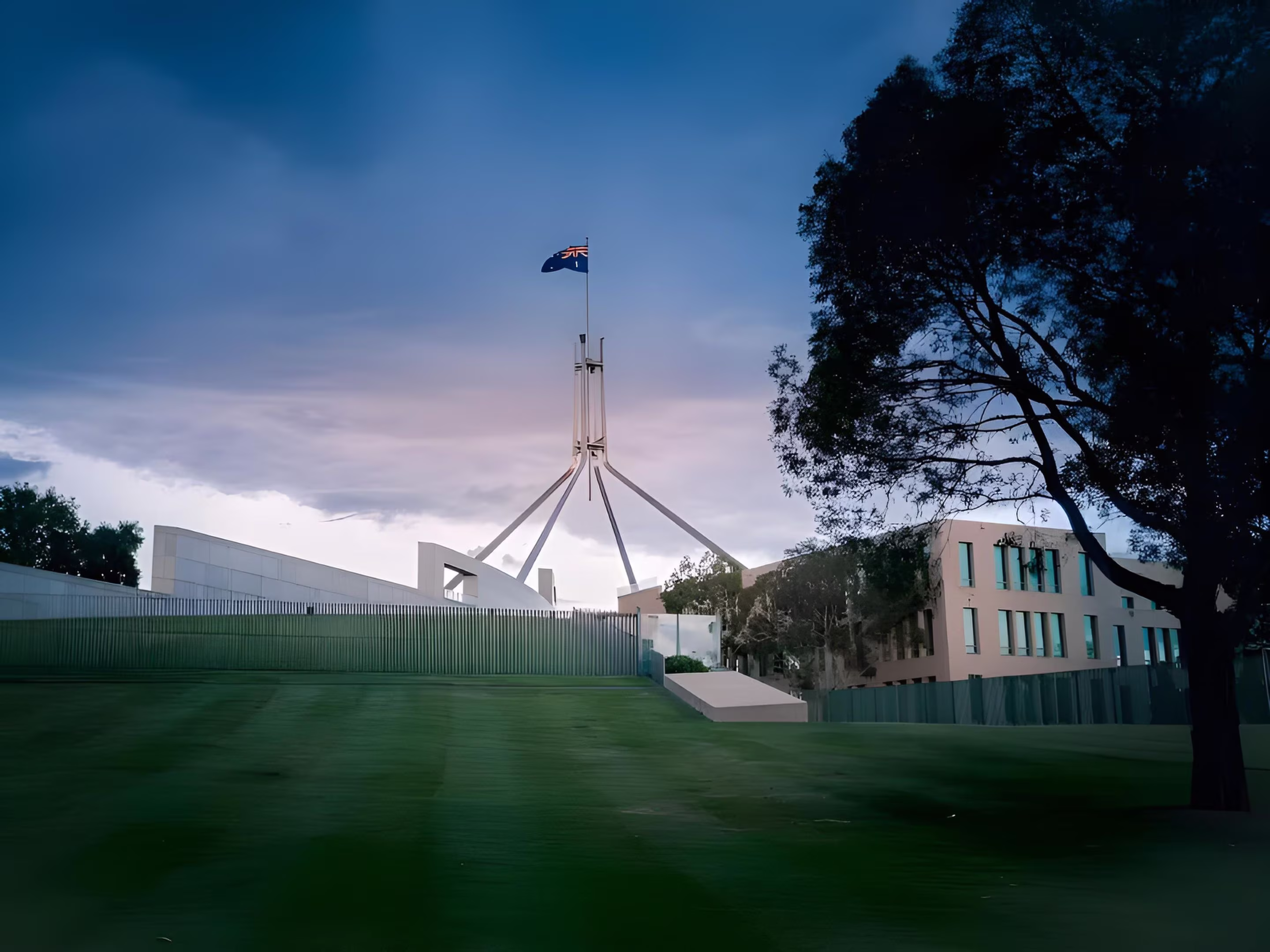 Australian Parliament House with an Australian flag flying on the flagpole against a cloudy sky at dusk.