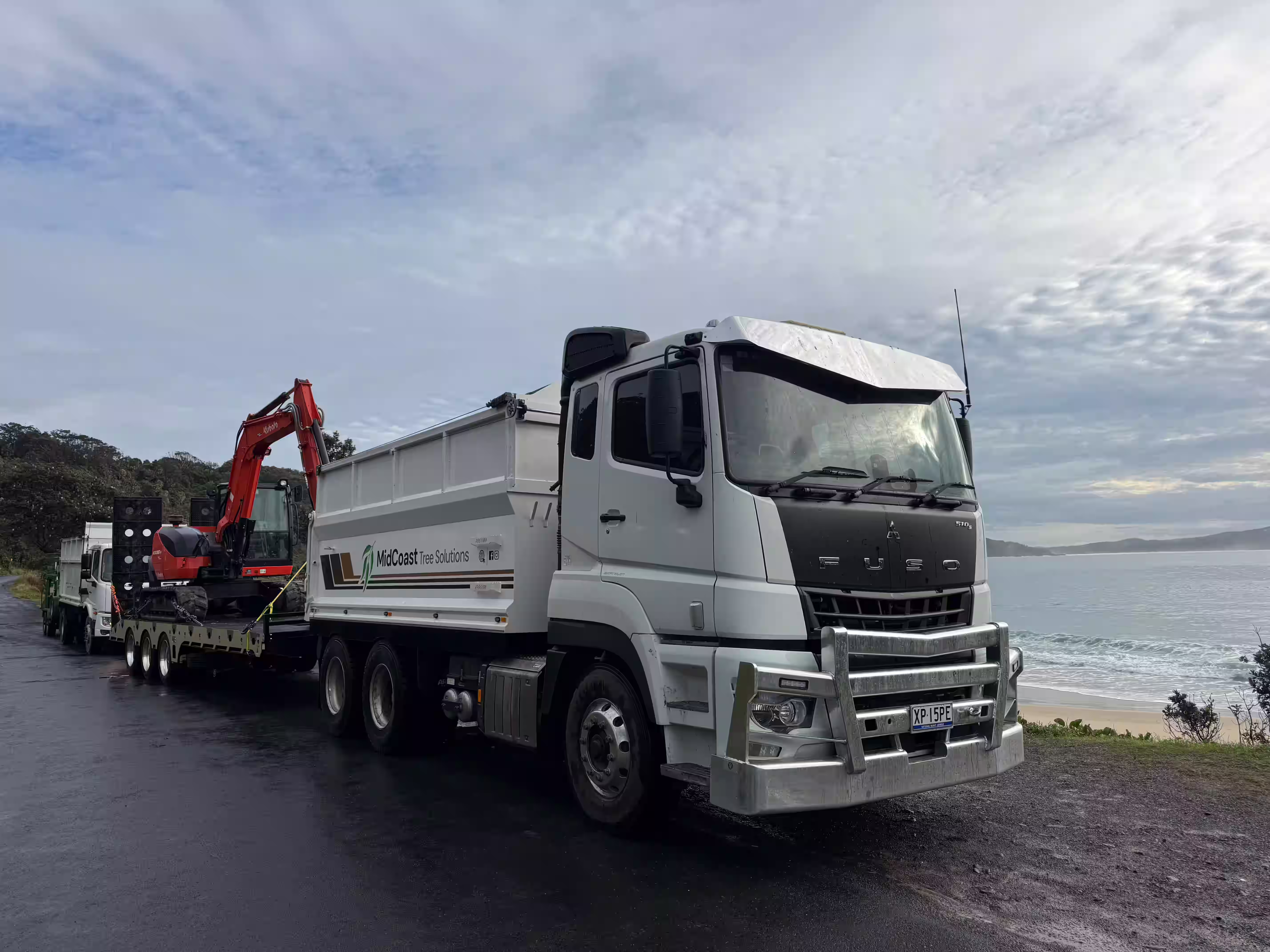 White Fuso truck with MidCoast Tree Solutions logo parked near a beach with an excavator on a trailer behind it.
