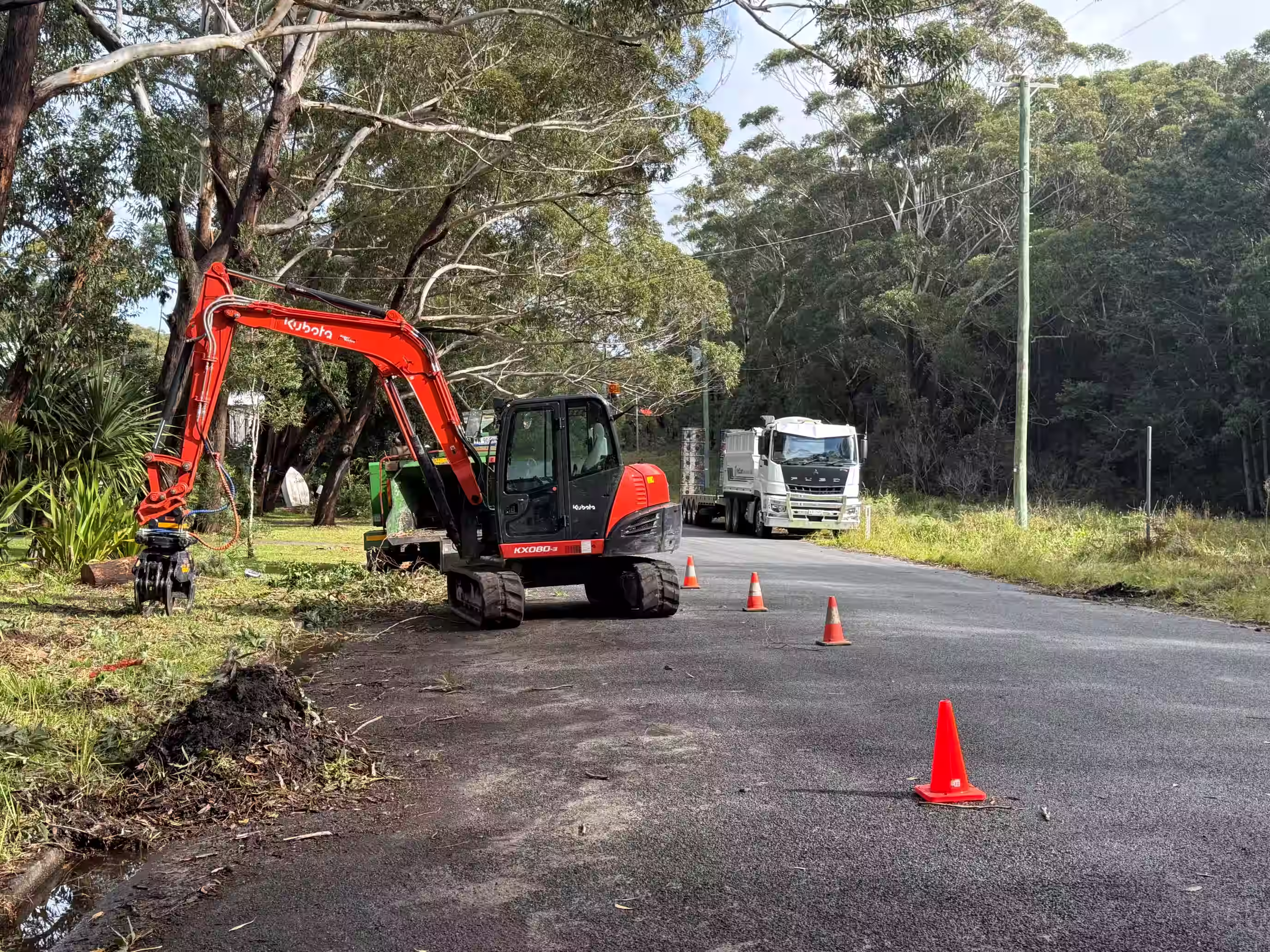 Red Kubota excavator on road edge near trees with traffic cones and a white truck in background.