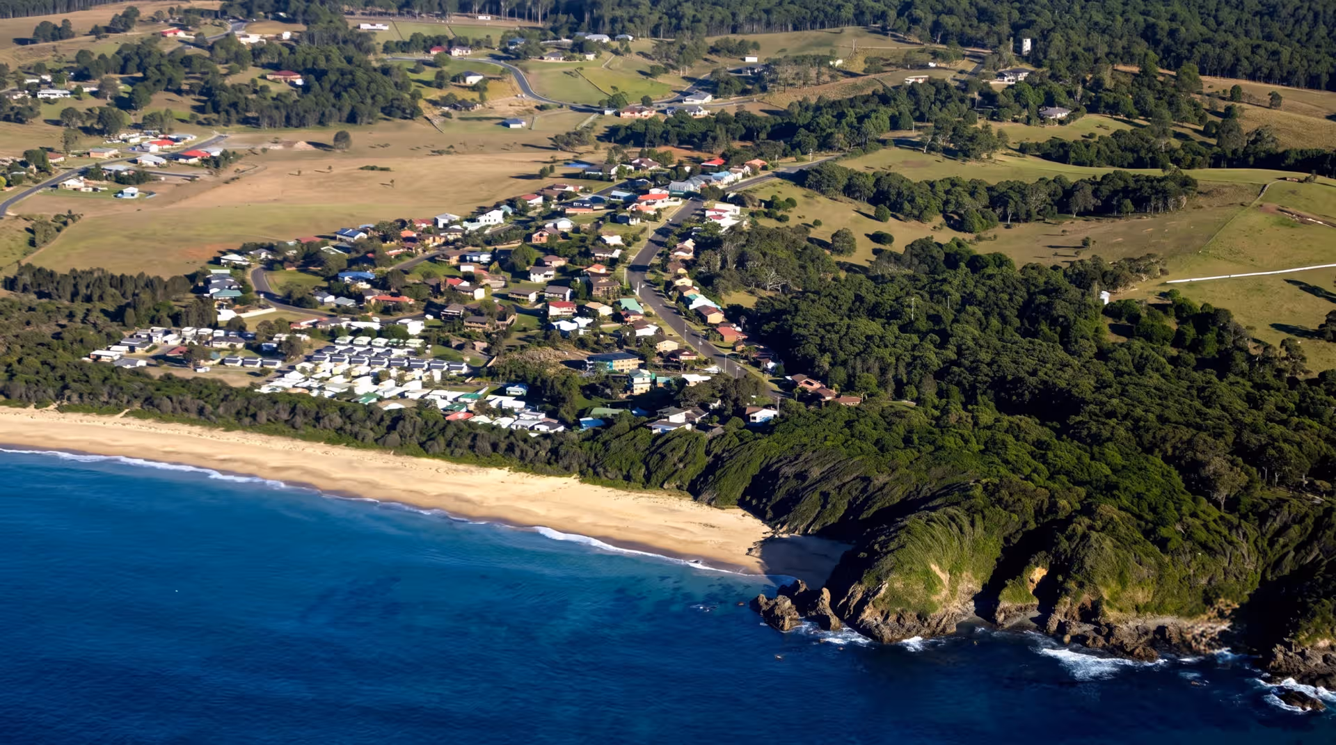 Aerial view of a coastal town with houses, sandy beach, forested cliffs, and blue ocean waters.