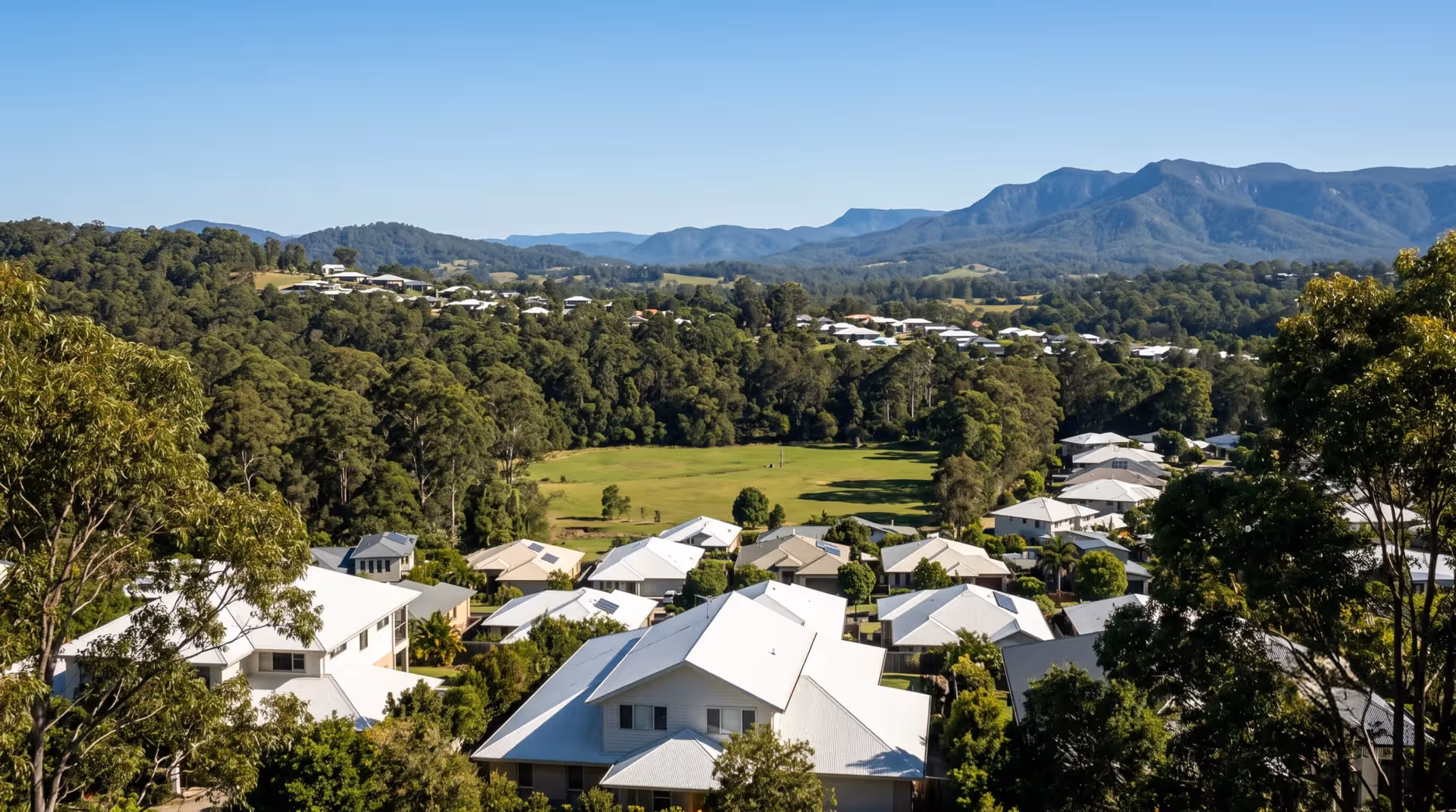A suburban neighborhood with white-roofed houses surrounded by trees and mountains under a clear blue sky.