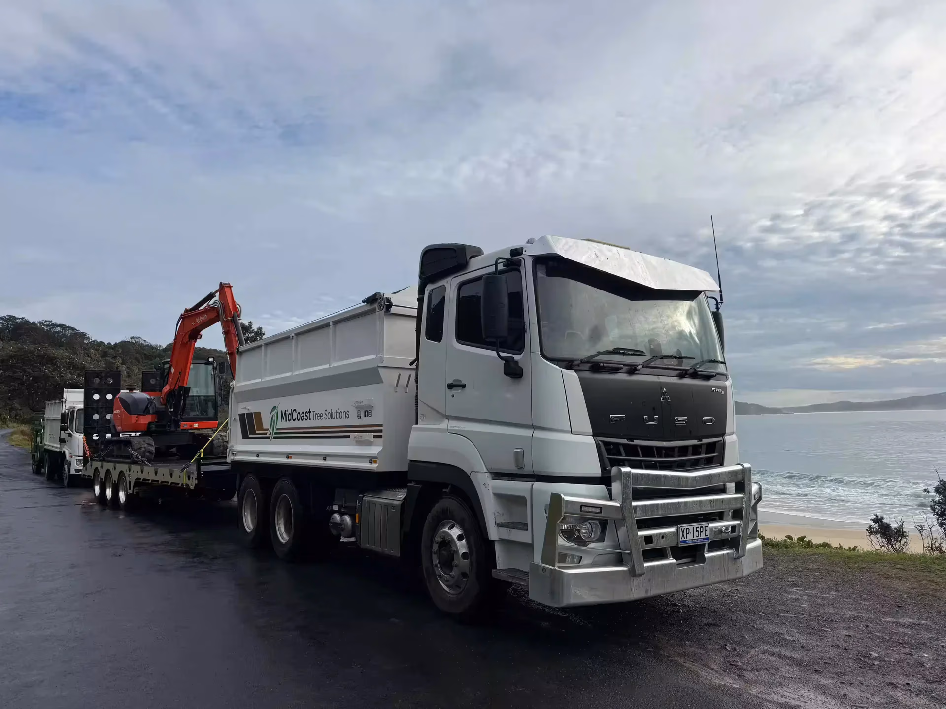 White MidCoast Tree Solutions truck with excavator on trailer parked by a coastal road.