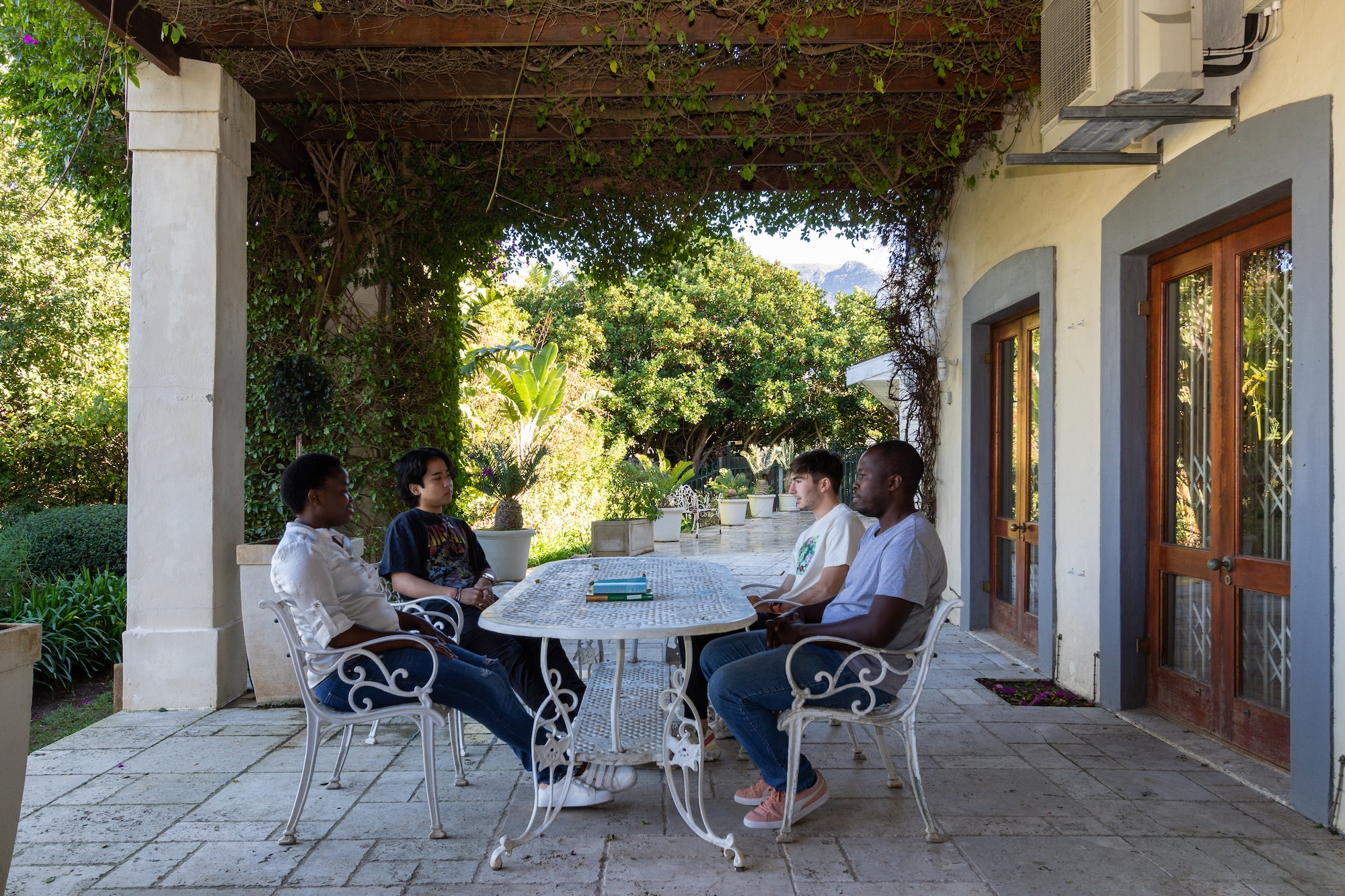 Bewoners in gesprek op het terras van de partnerkliniek in Zuid-Afrika Buitenaanzicht van een villa met palmbomen en terras, een van de locaties van Affect2U in het buitenland.