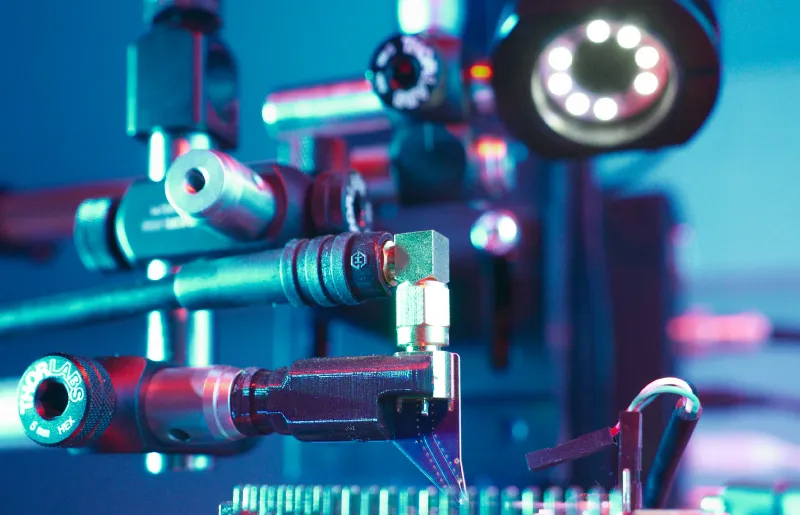 Close-up of a scientific EMFI optical setup with lenses, mirrors, and electronic components on a laboratory bench under blue and purple lighting.