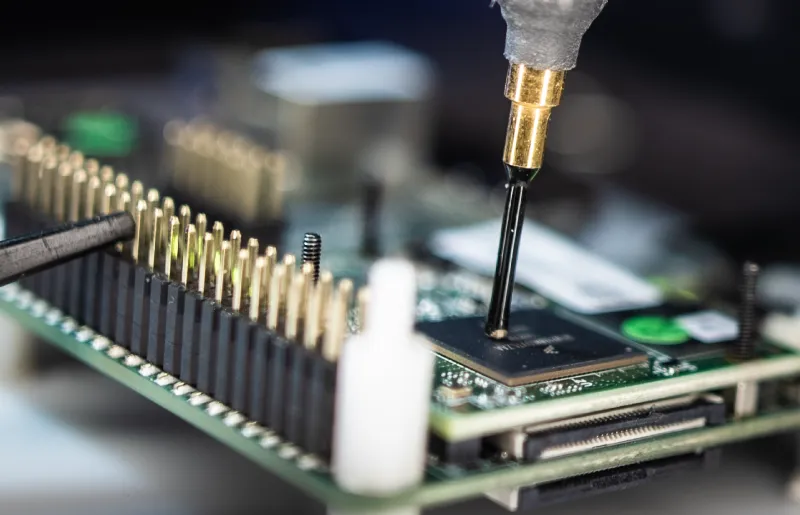 Close-up of a SCA Near-Field EM Leakage Probes gold-tipped probe touching a black microchip mounted on a green circuit board with multiple gold pins.