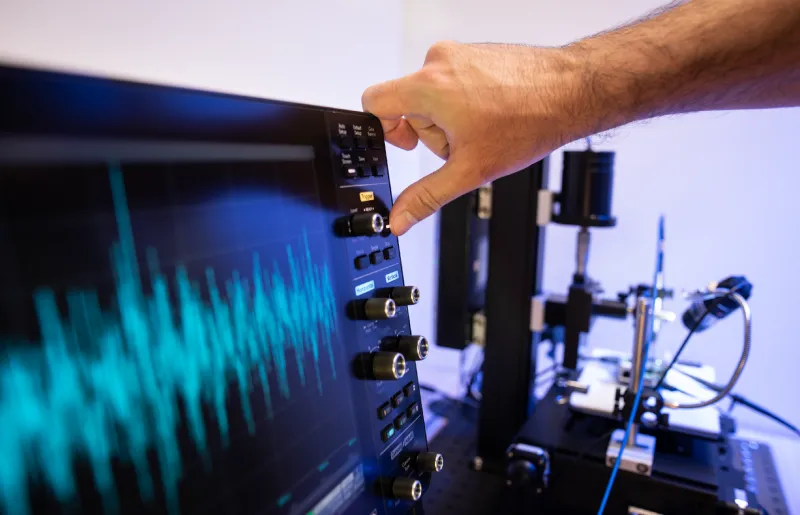 Hand adjusting a knob on an oscilloscope displaying a blue waveform in a laboratory setting with scientific equipment in the background.