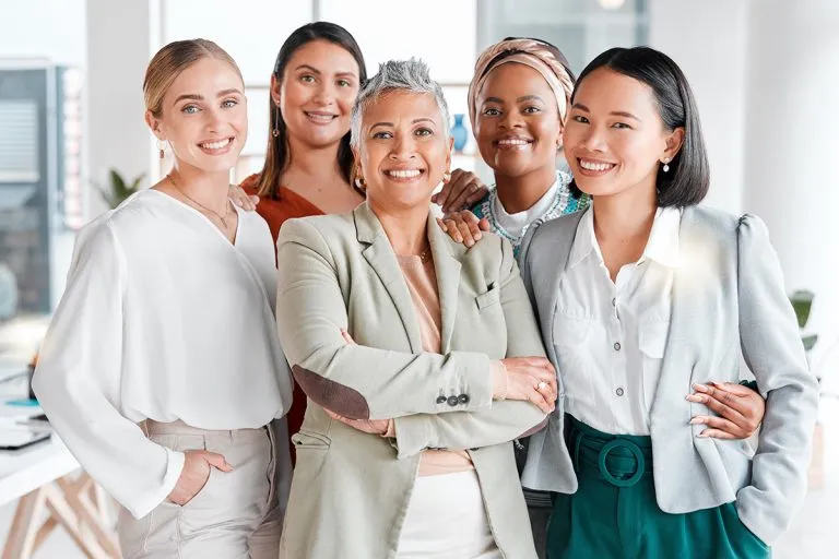 Group of five diverse professional women smiling and standing closely together in a bright office.