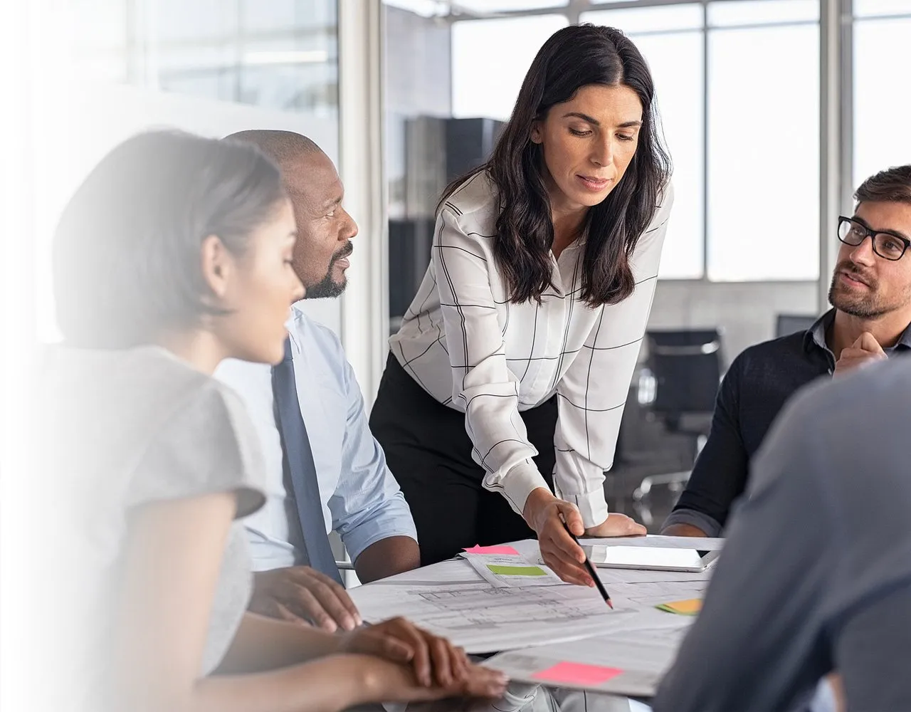 Businesswoman leading a meeting while pointing at documents spread on a table with colleagues attentively listening.