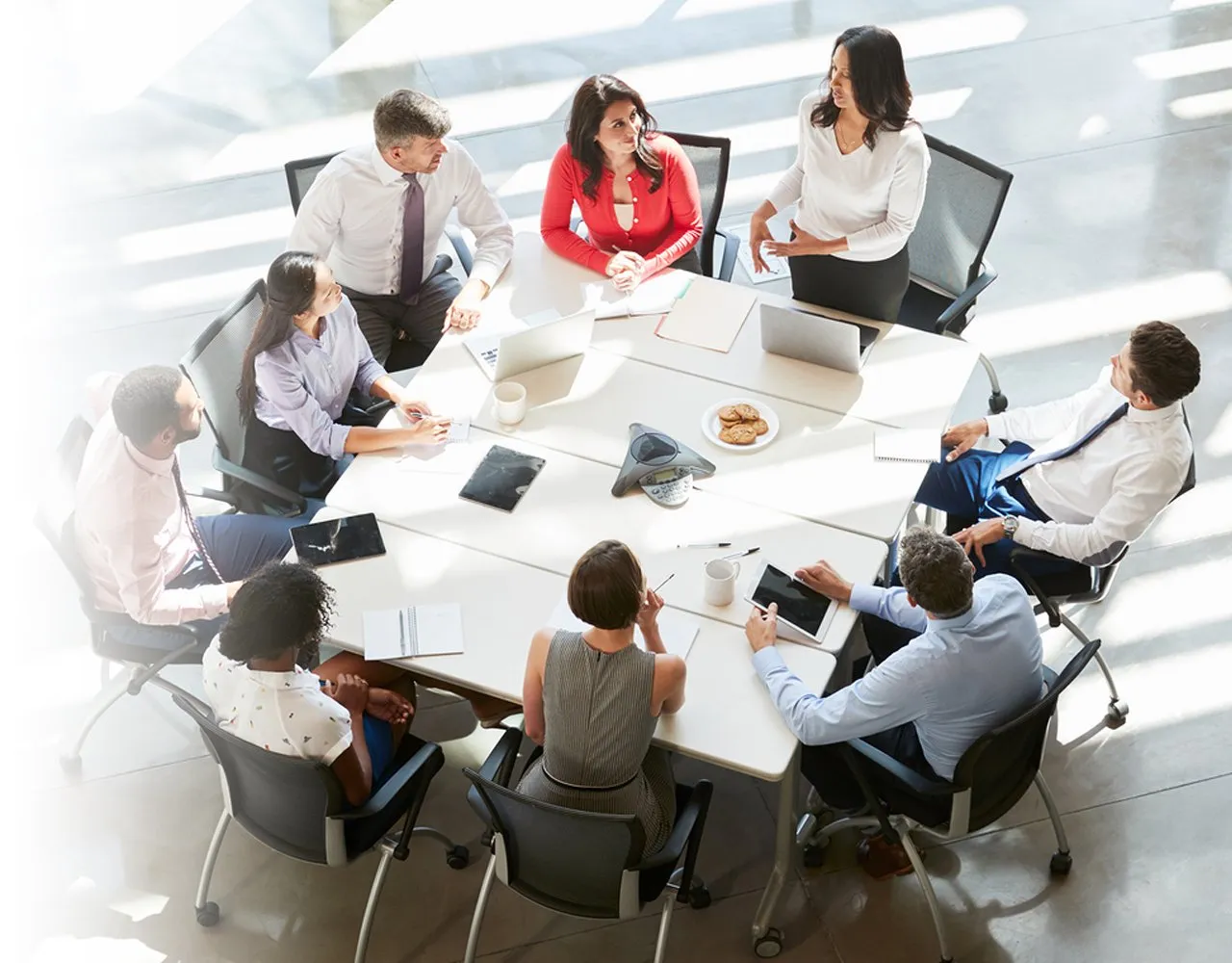 Eight diverse professionals in business attire having a meeting around a white conference table with laptops, tablets, notebooks, and a plate of cookies.